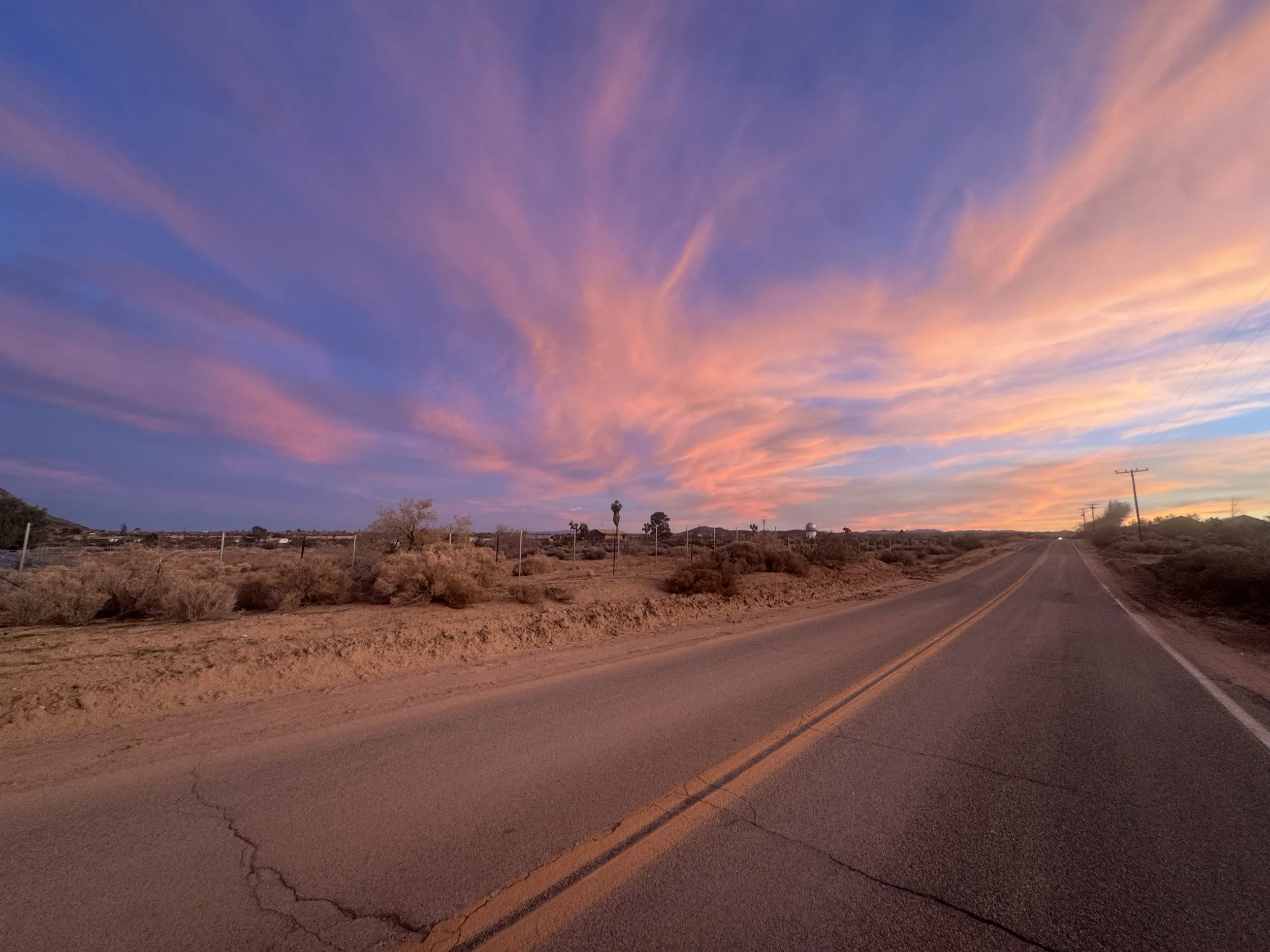 A desolate road running through a desert landscape during sunset, with colorful clouds in the sky and utility poles along the side.
