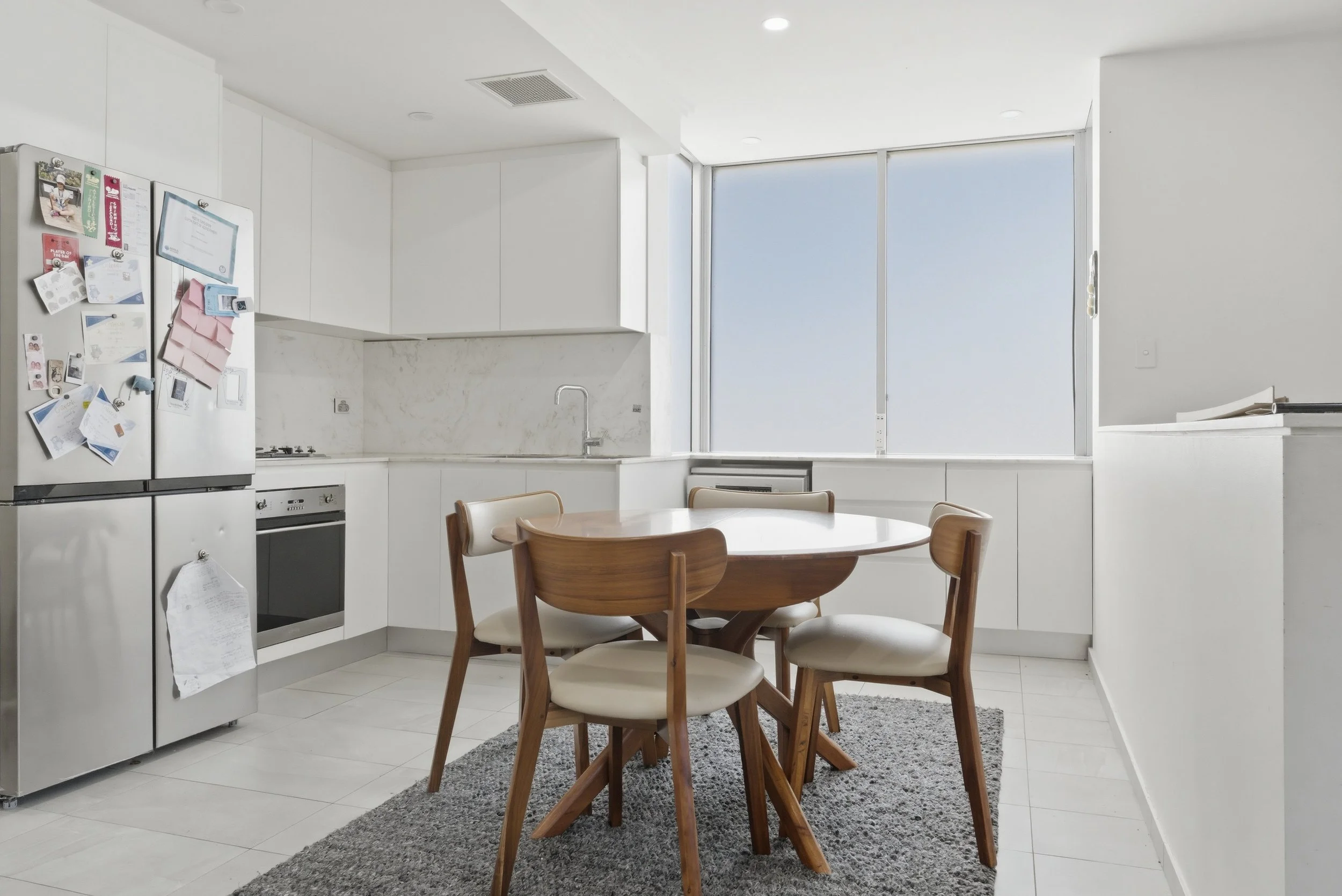 A modern, bright kitchen with white cabinets and walls, a silver refrigerator covered with magnets and papers, a small gas stove, a sink, large windows with shades, and a round wooden dining table with chairs on a gray rug.