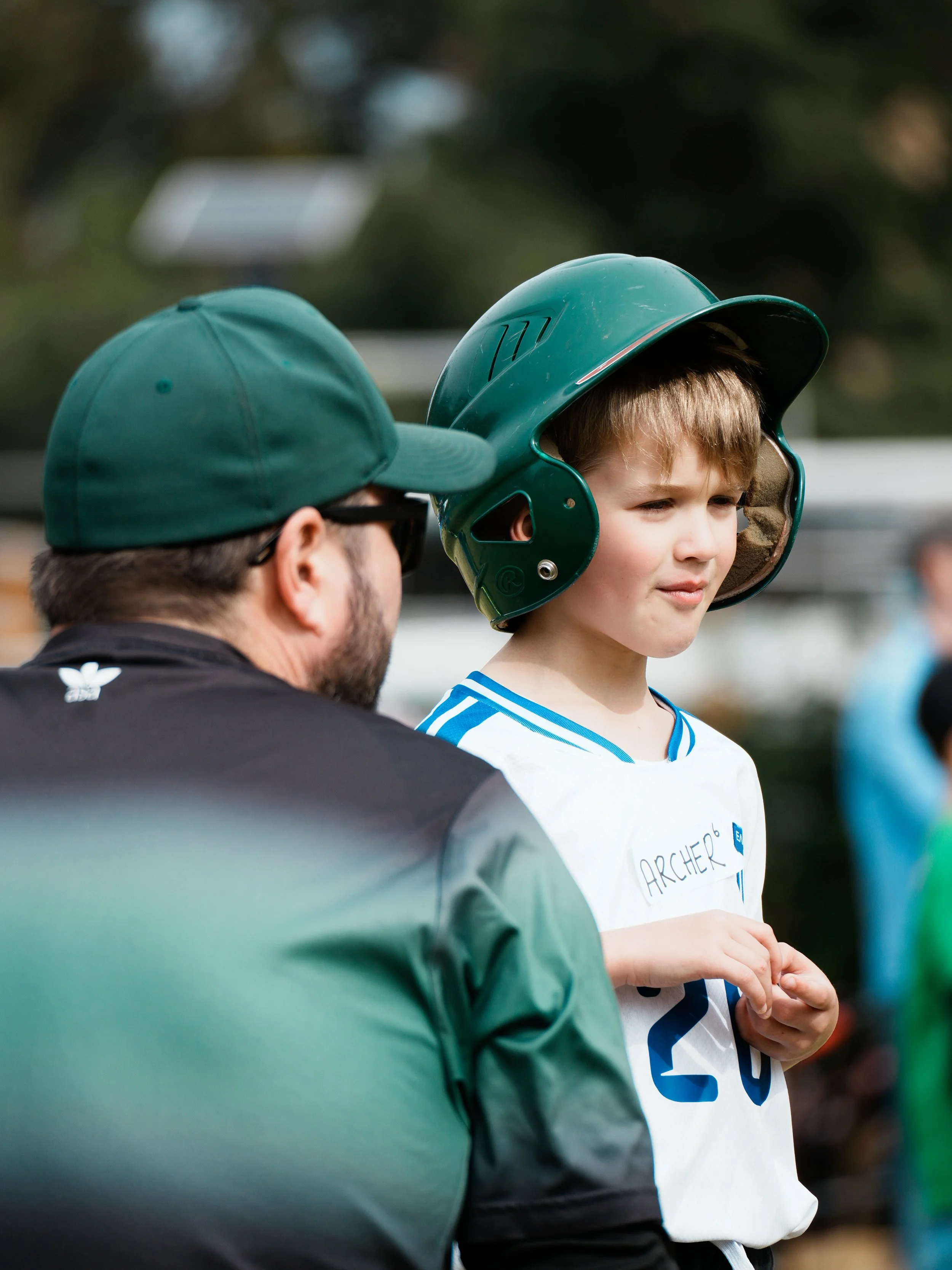 A young boy with short blonde hair wearing a white shirt with blue and black accents and a name tag that reads 'Archers' is wearing a protective green baseball helmet with a face shield attached. An adult man with glasses and a green cap is standing 