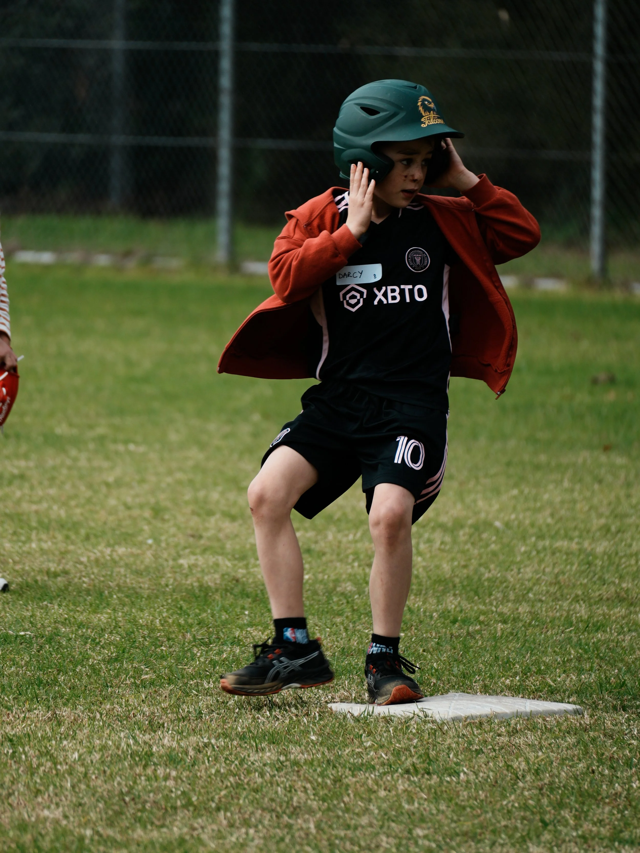 A young boy wearing a helmet, black sports uniform with the number 10, and a red jacket is preparing to bat at a baseball field. He is standing on a base with one foot on it, and holding his helmet with both hands while looking down.