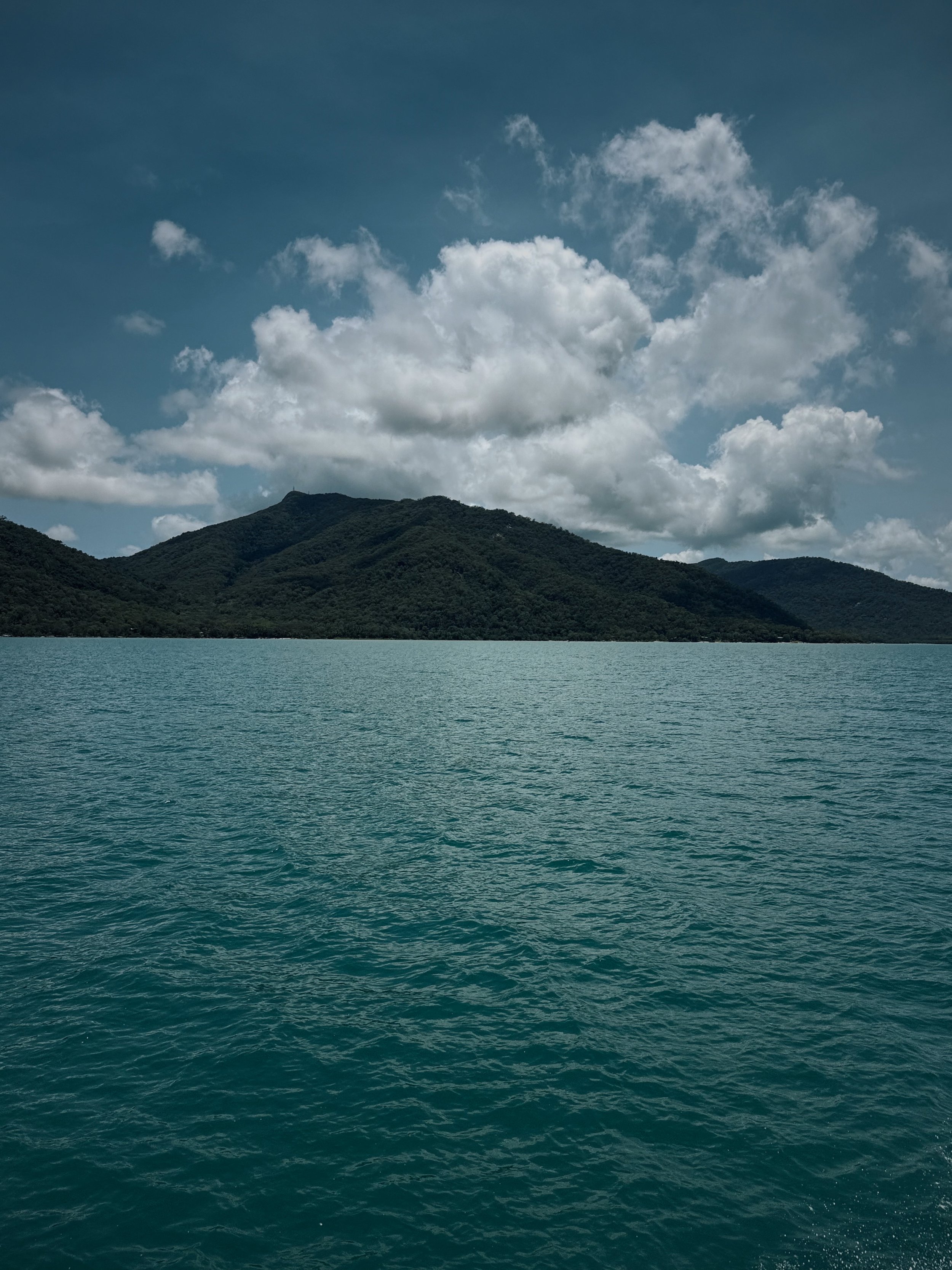 A lake with turquoise water in front of dark green mountains under a partly cloudy sky.