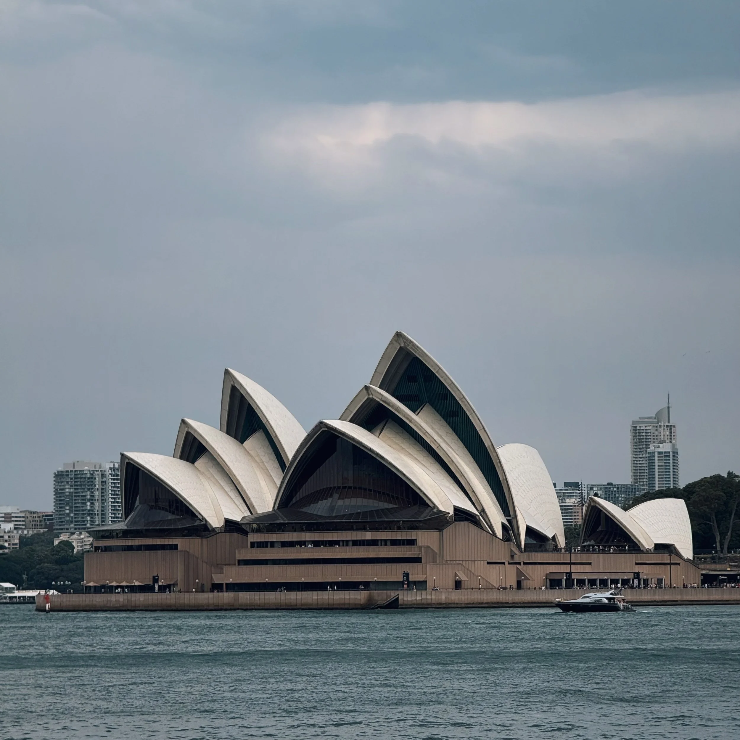 Sydney Opera House on the water under a cloudy sky, with nearby city buildings in the background.