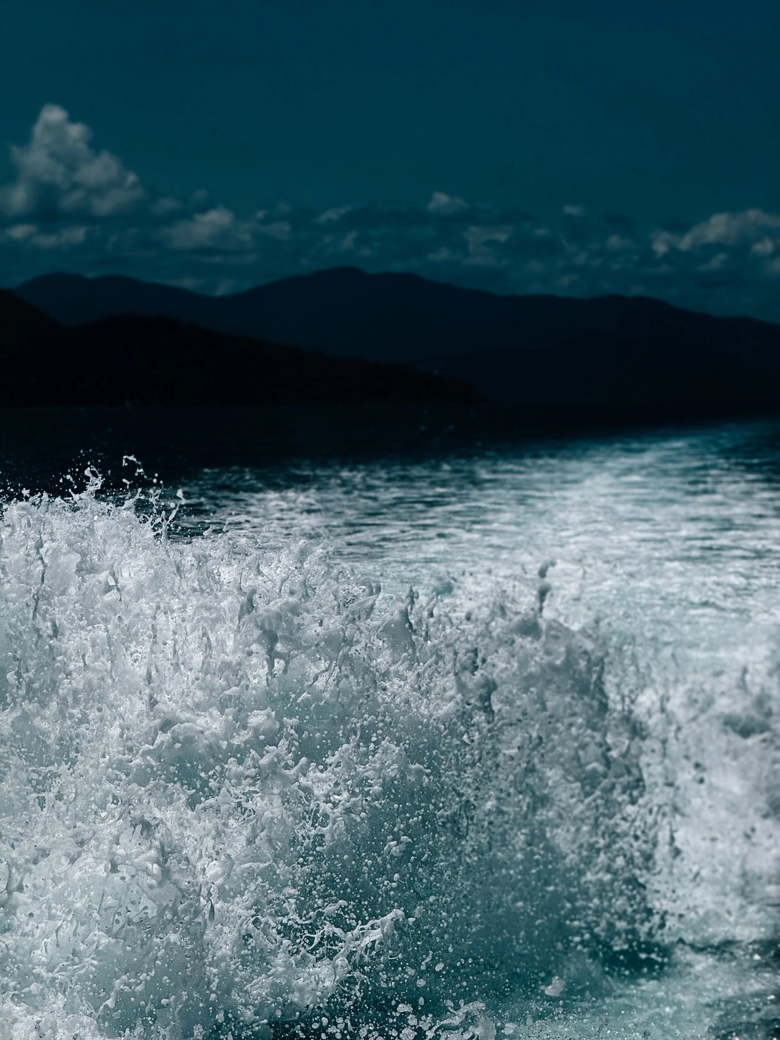 Waves crashing in the ocean with a silhouette of mountains and clouds in the background under a dark sky.