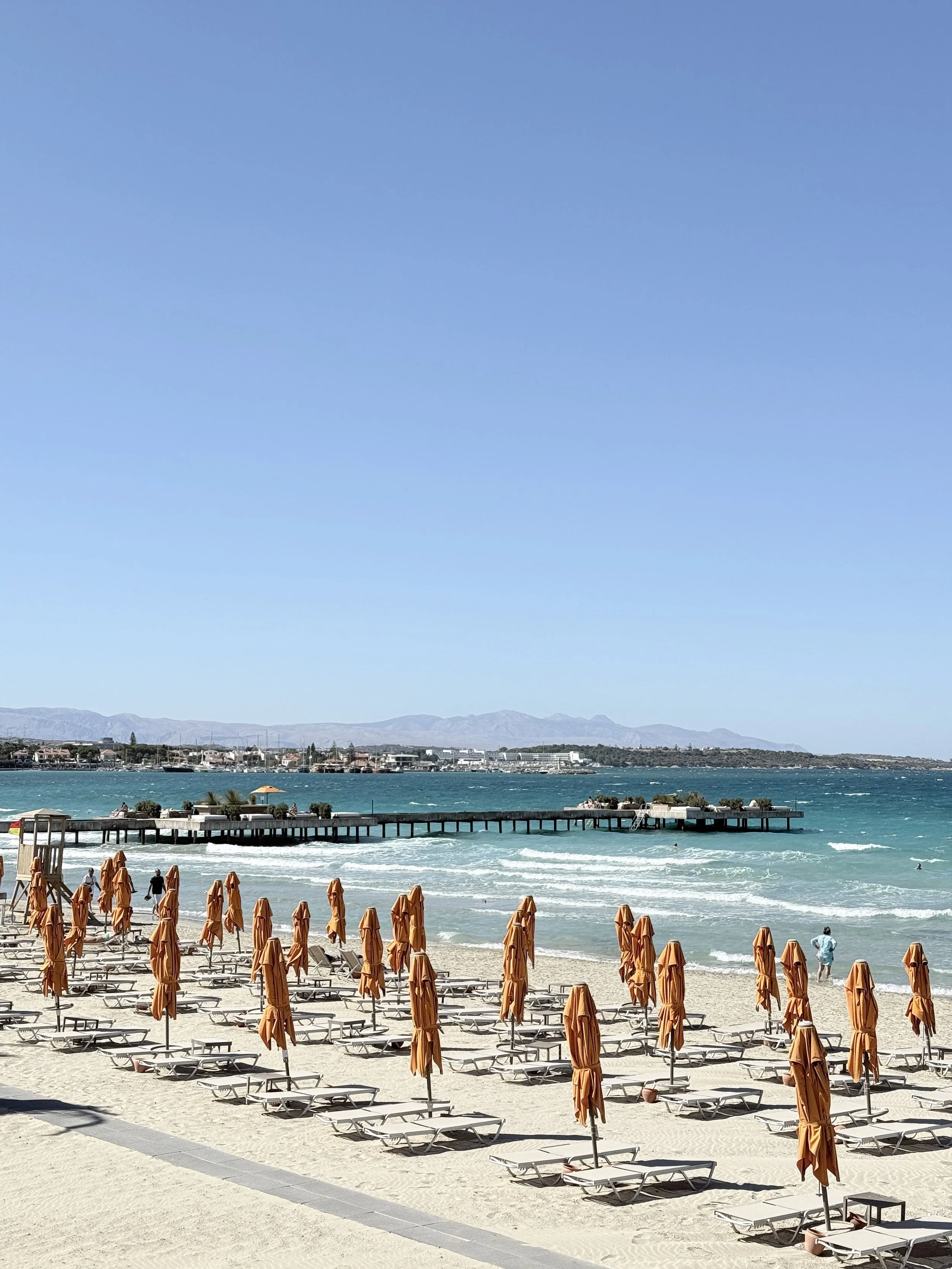 A beach with orange umbrellas and lounge chairs along the sand, a pier extending into the water, and mountains in the background under a clear blue sky.