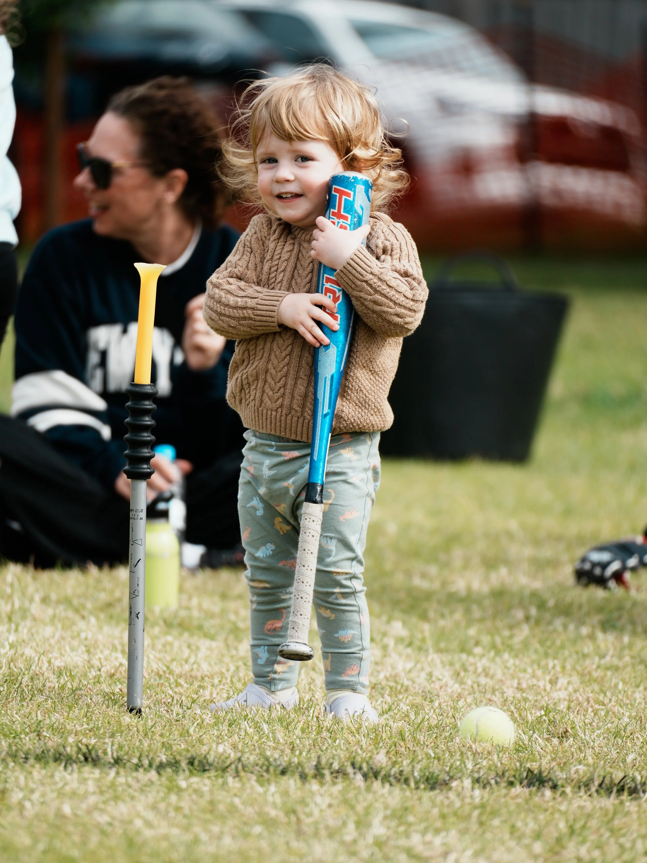 Young girl with red curly hair holding a hockey stick on a grassy field, with a tennis ball nearby, and a woman with dark hair and sunglasses sitting in the background.