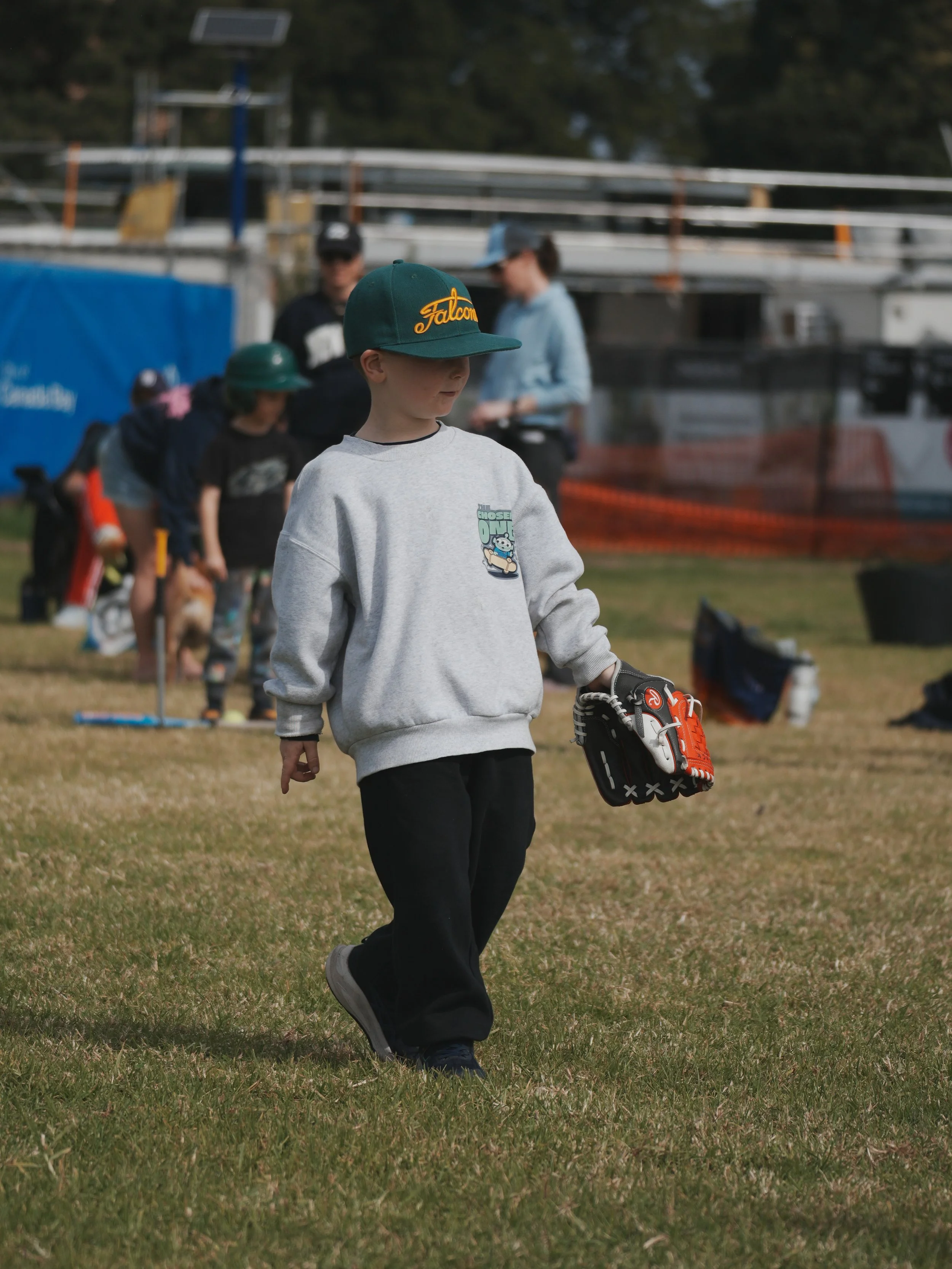 A young boy in a gray sweatshirt and black pants walking on a grassy field holding a baseball glove, with several children and adults in the background near a sports field.