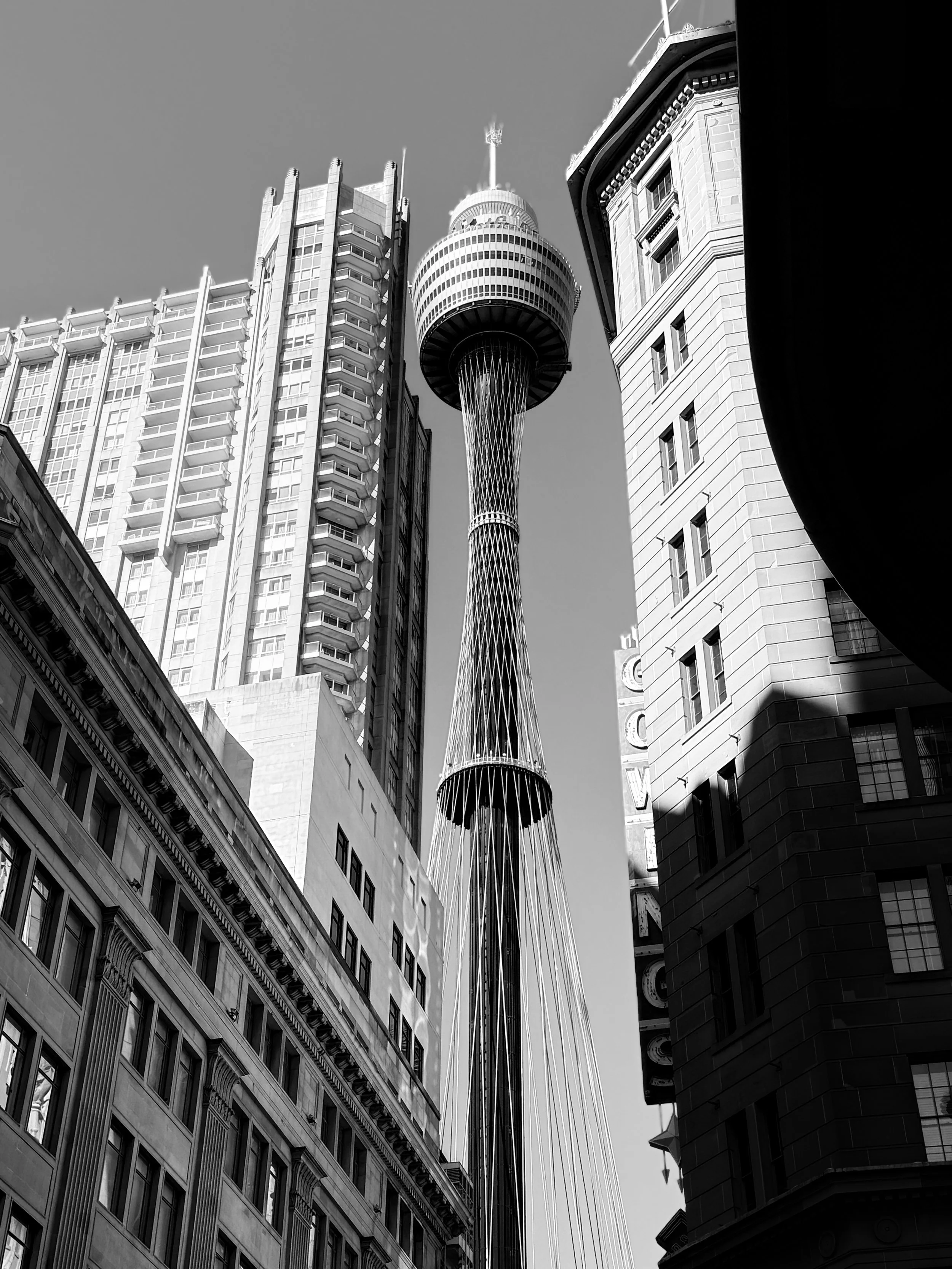 Black and white photo of a tall tower with a circular observation deck, surrounding buildings with multiple floors and architectural details, viewed from a narrow street.