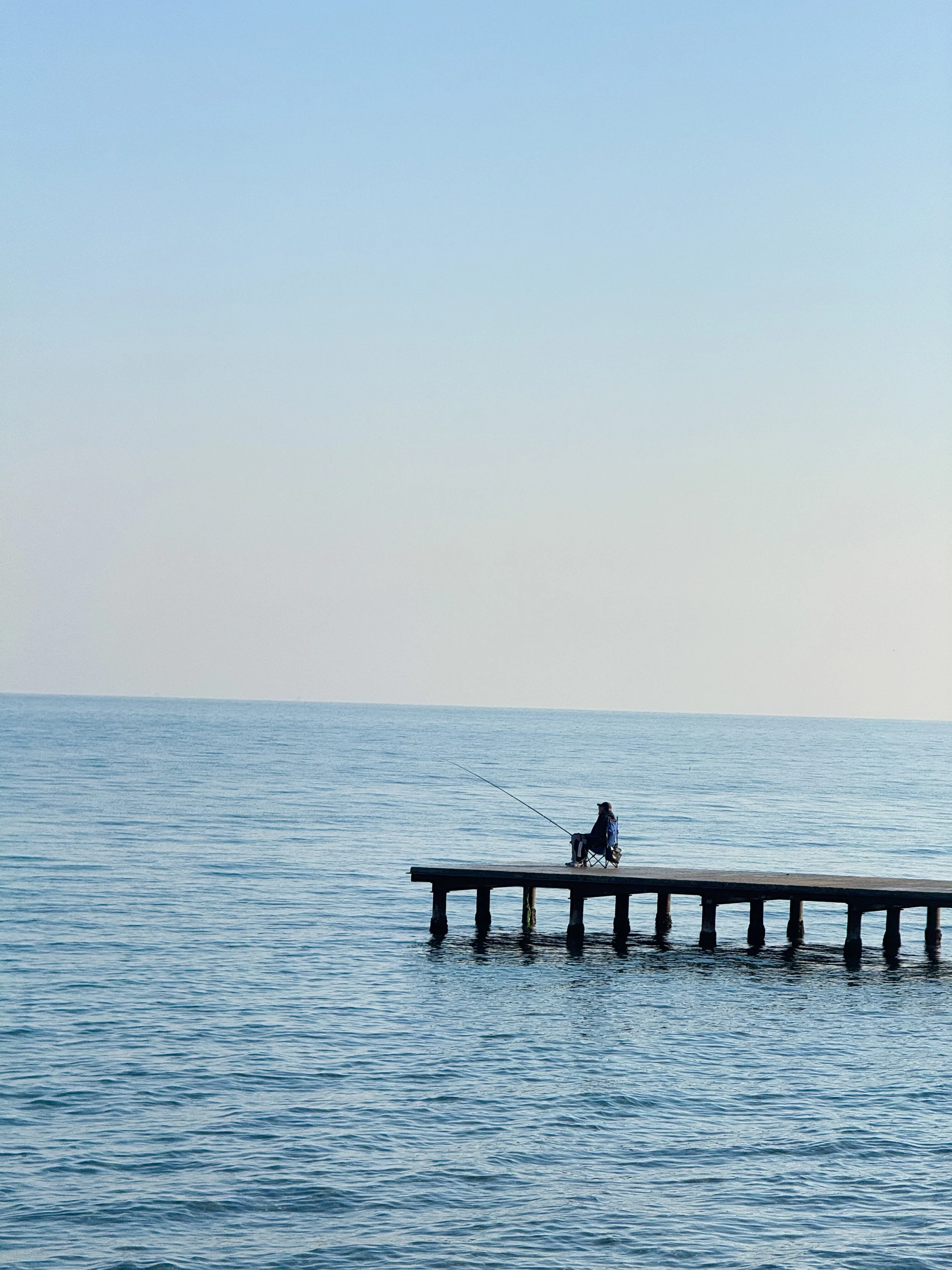 A person sitting on a dock fishing in the ocean during daylight.