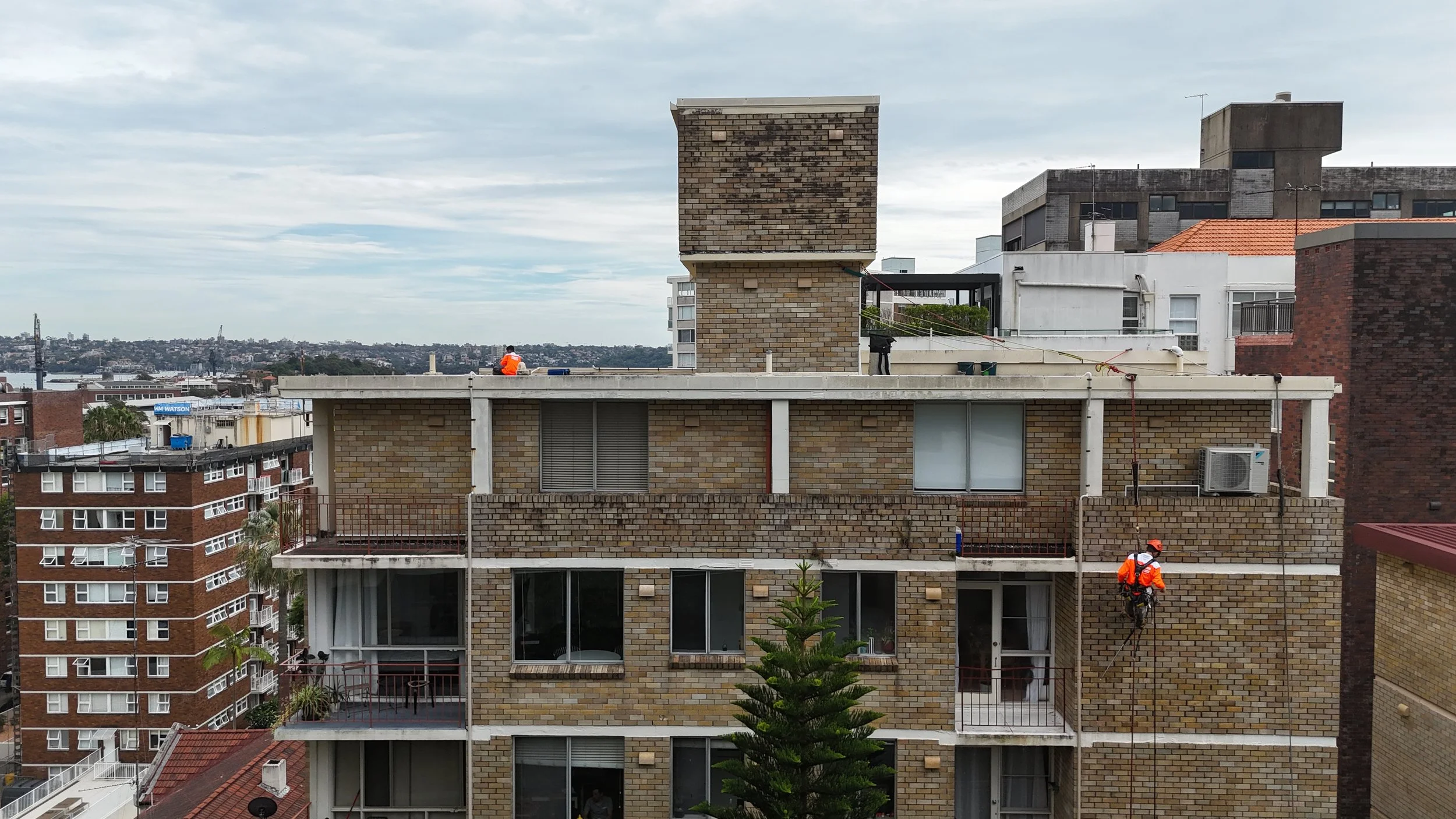Building workers in orange safety gear performing maintenance on the exterior of a multi-story brick apartment building.