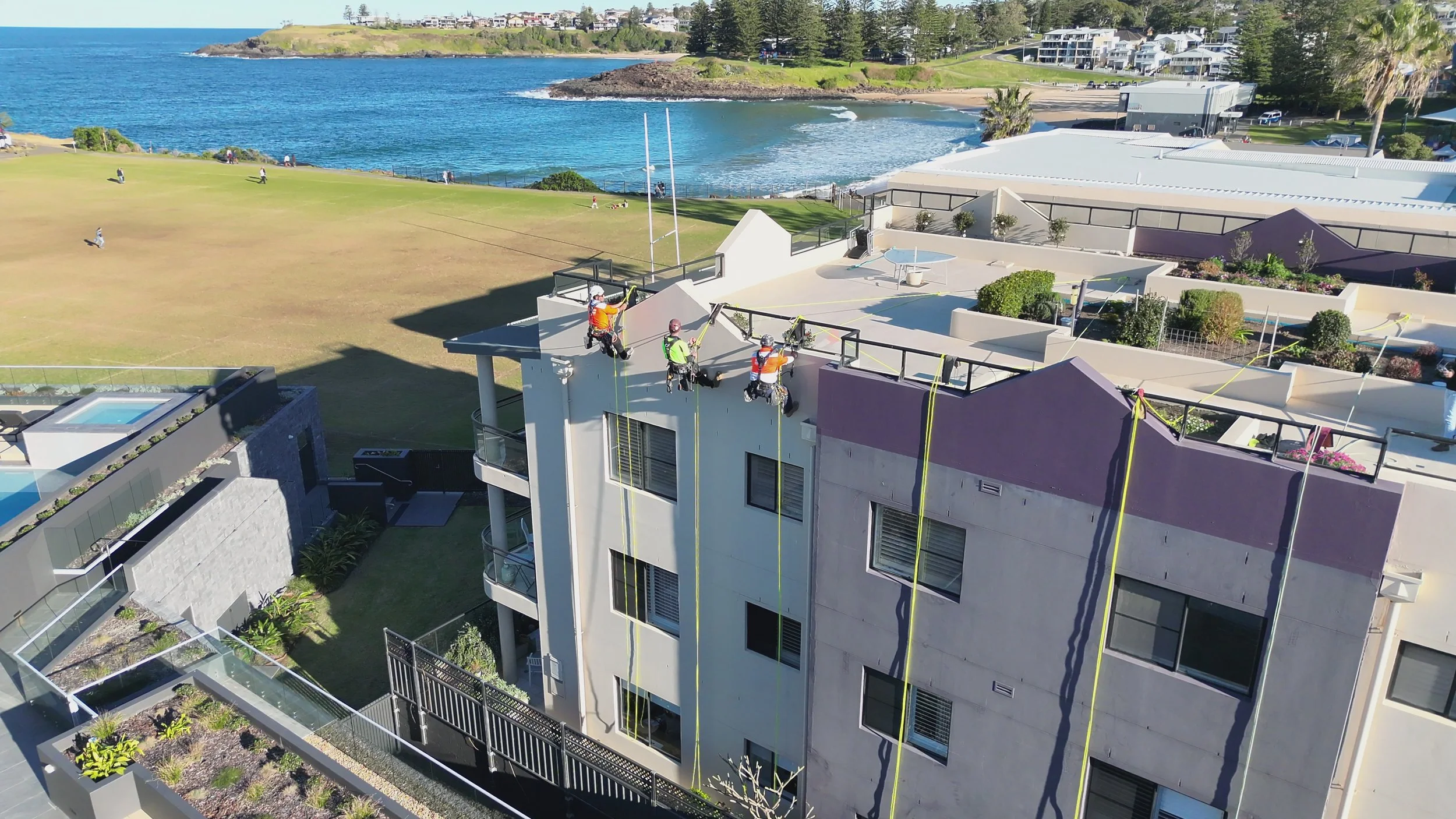 Construction workers wearing safety harnesses working on the exterior of a multi-story building near a coastal area, with grassy fields and the ocean in the background.