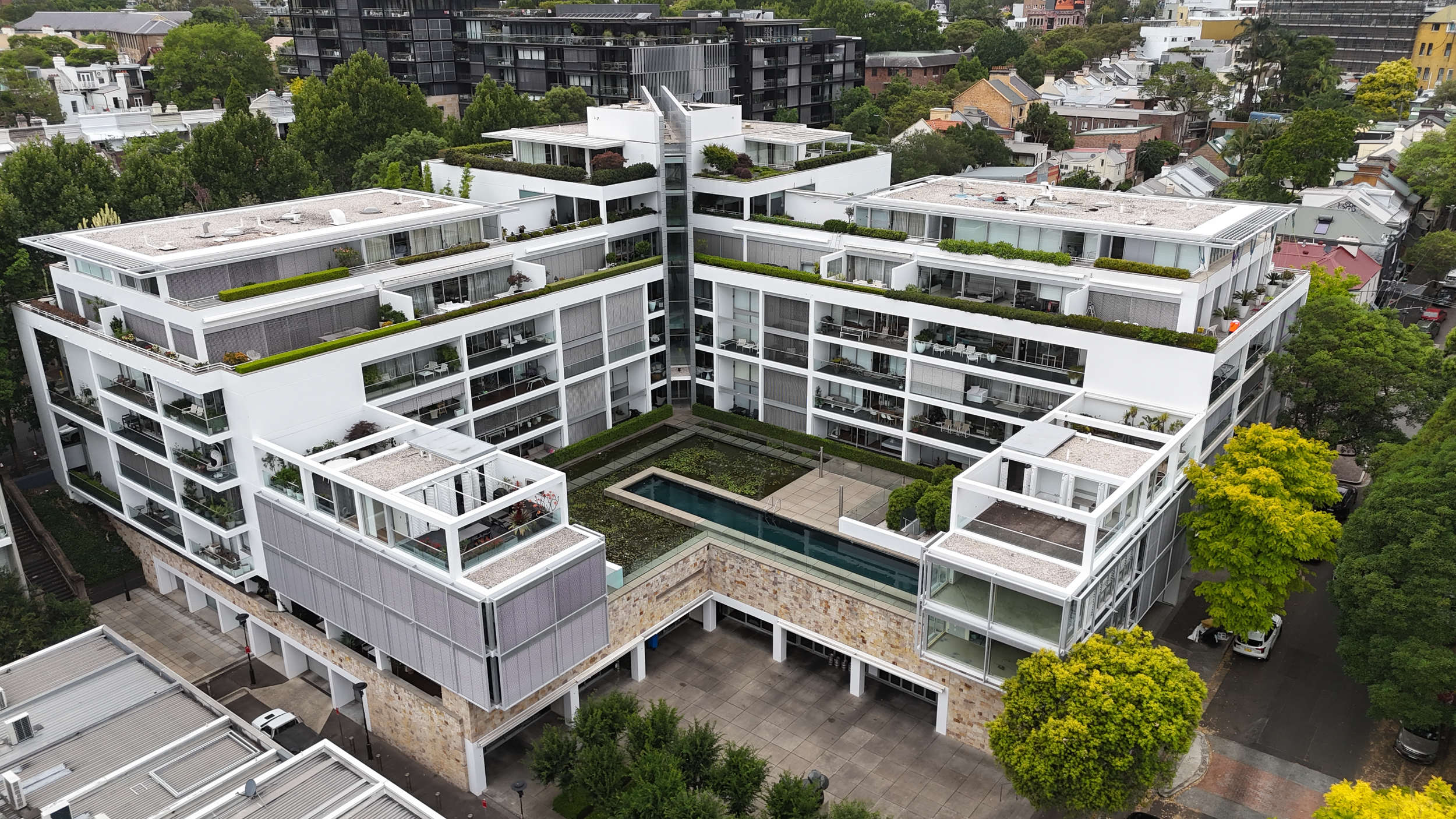 A modern, white, multi-story apartment building with terraces and rooftop gardens, featuring a central courtyard with a swimming pool, surrounded by trees and neighboring buildings.