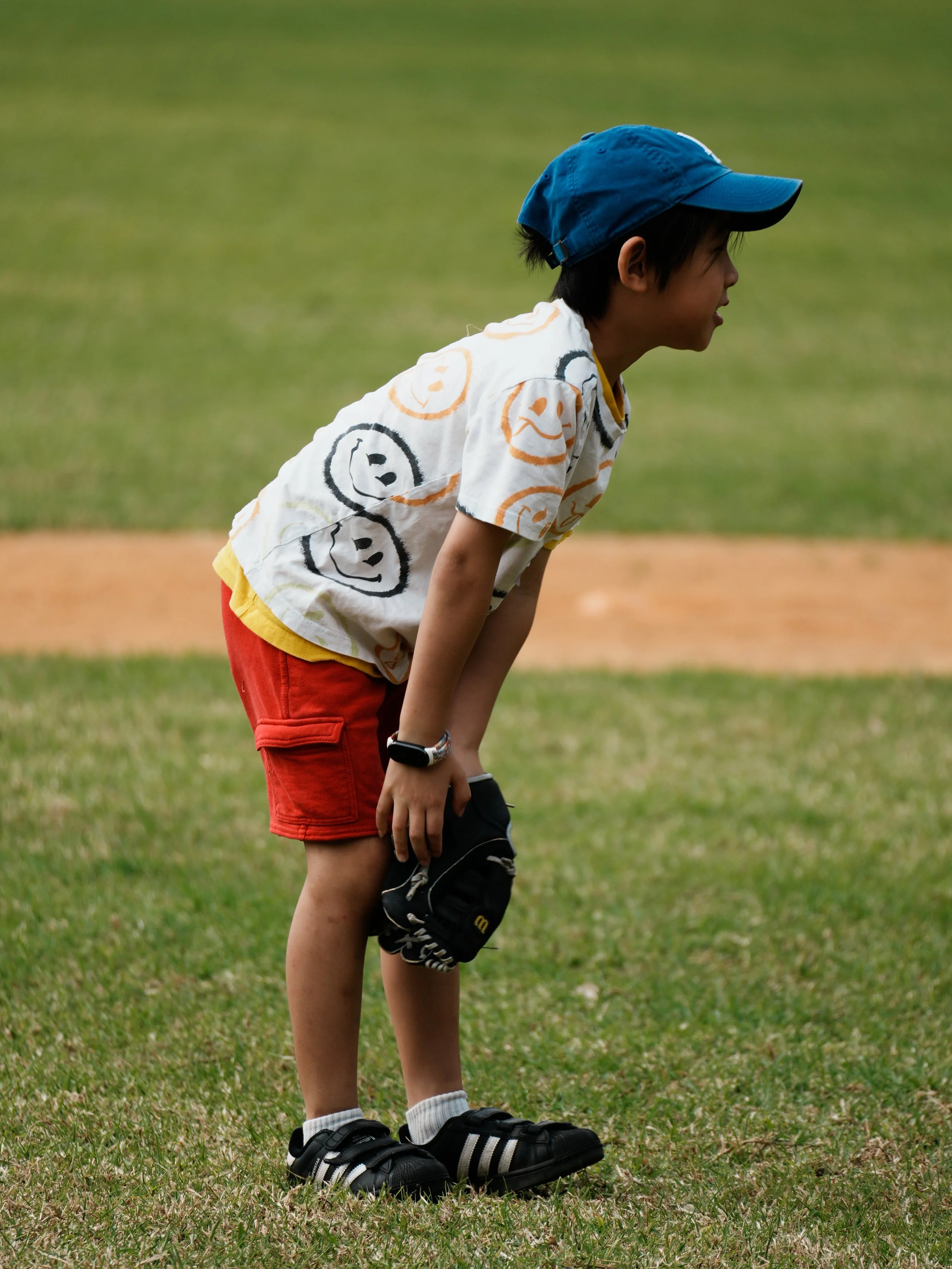 A young boy with dark hair wearing a blue cap, a white T-shirt with orange and black smiley face prints, red shorts, white socks, and black Nike sneakers stands on a grassy field, holding a black cap in his hand, appearing to be in a sports or outdoo