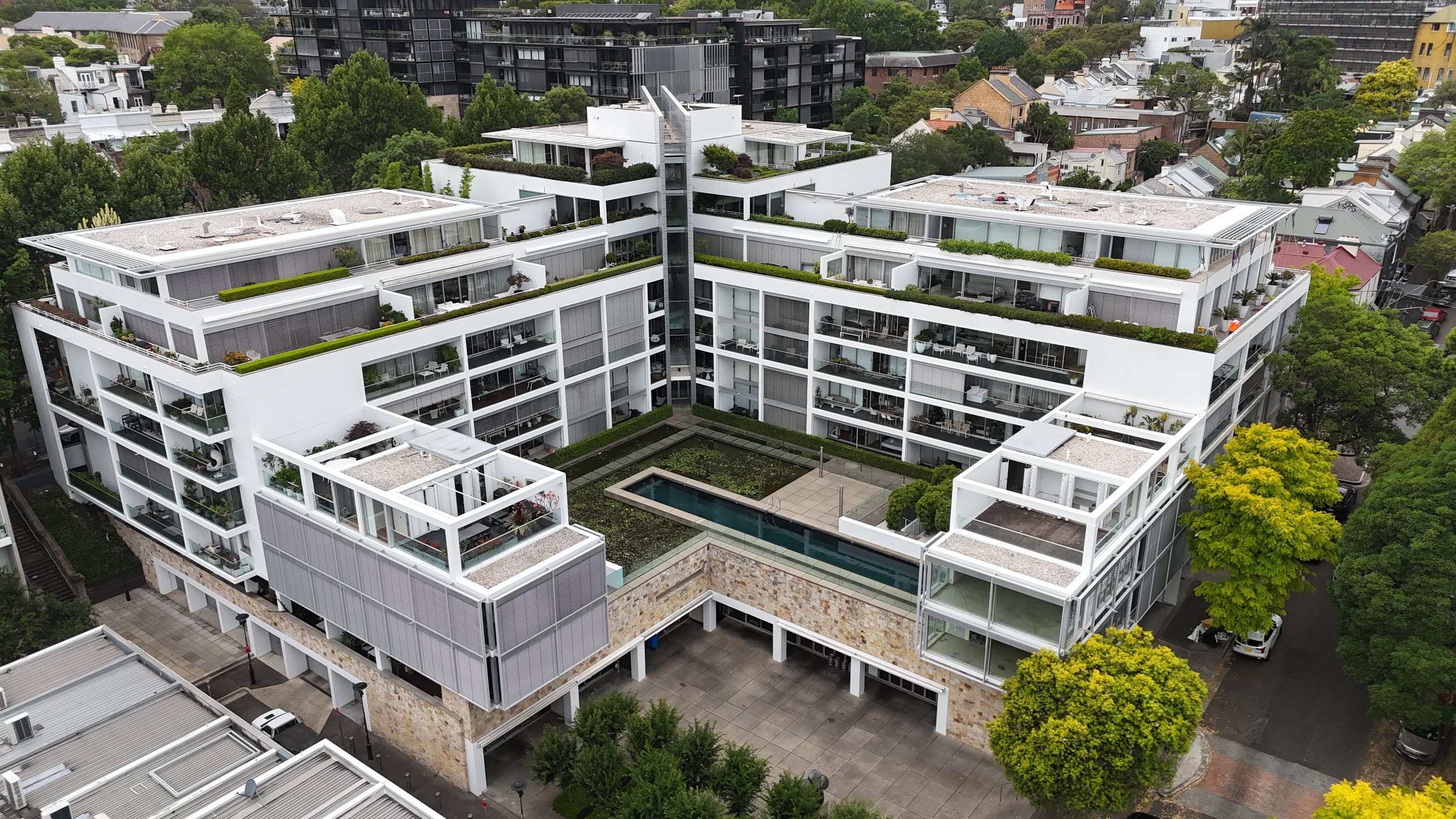 Aerial view of a modern white apartment building with rooftop gardens, a small pool, and surrounding trees in an urban neighborhood.