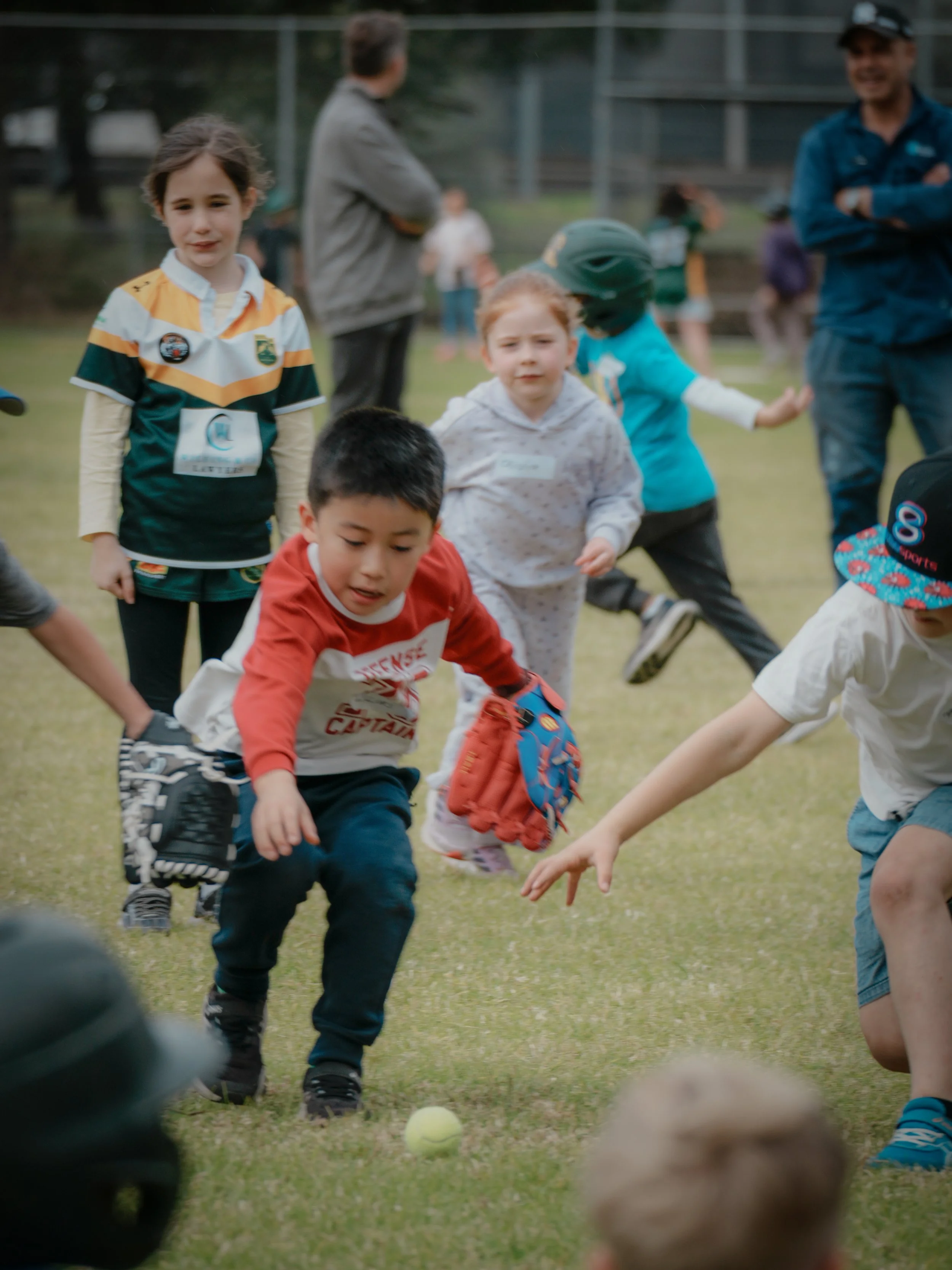 Children playing baseball on a grassy field with adults watching in the background.