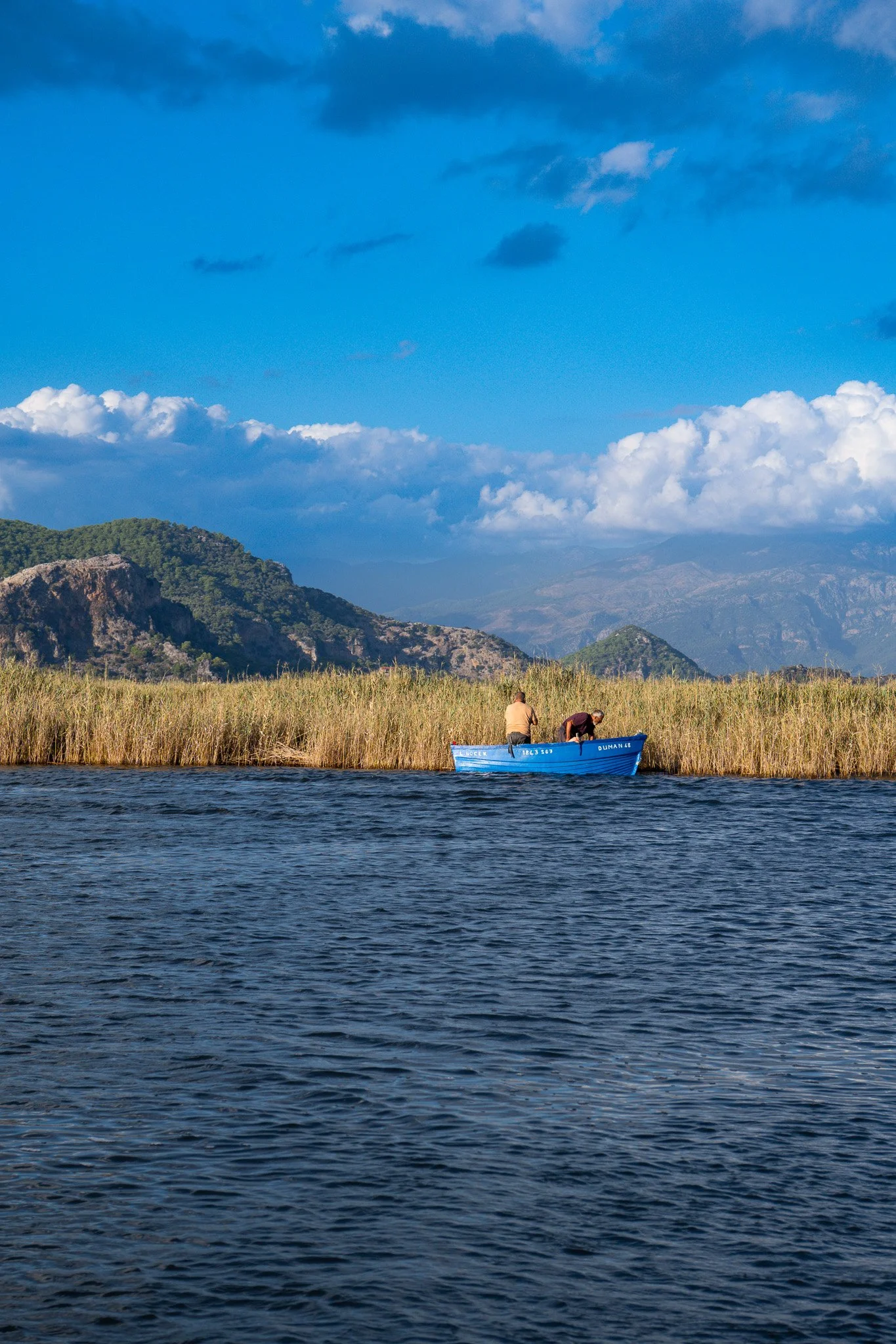 Two people in a small blue boat on a body of water, with tall grass and mountains in the background under a partly cloudy sky.