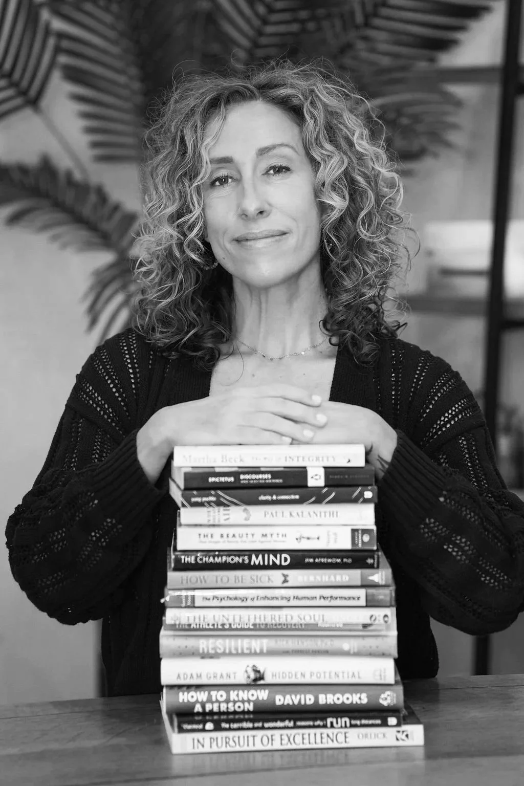 black and white image of a therapist smiling and sitting at her desk with hands resting on a large stack of books
