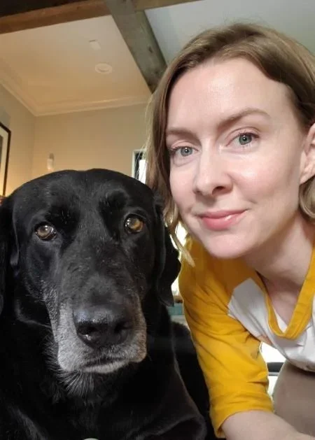 A woman with shoulder-length light brown hair taking a selfie with a black Labrador retriever dog, both indoors near a window with natural light.