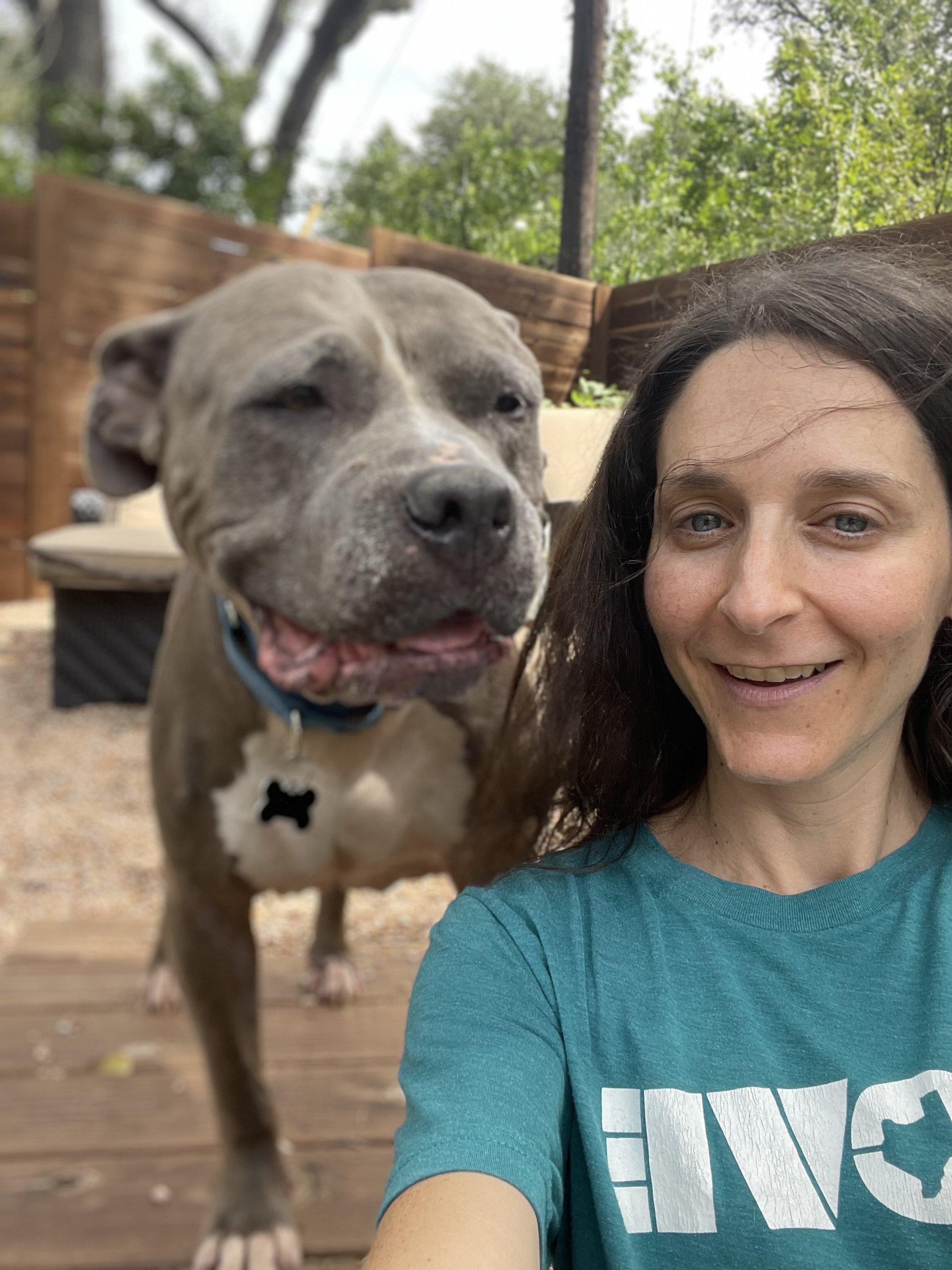 A woman, Katya Lidsky, taking a selfie with a happy, gray, brindle pitbull rescue dog outdoors in a backyard with trees and a wooden fence.