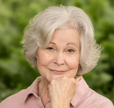 An elderly woman with gray hair smiling softly, resting her chin on her hand, outdoors with green foliage in the background.
