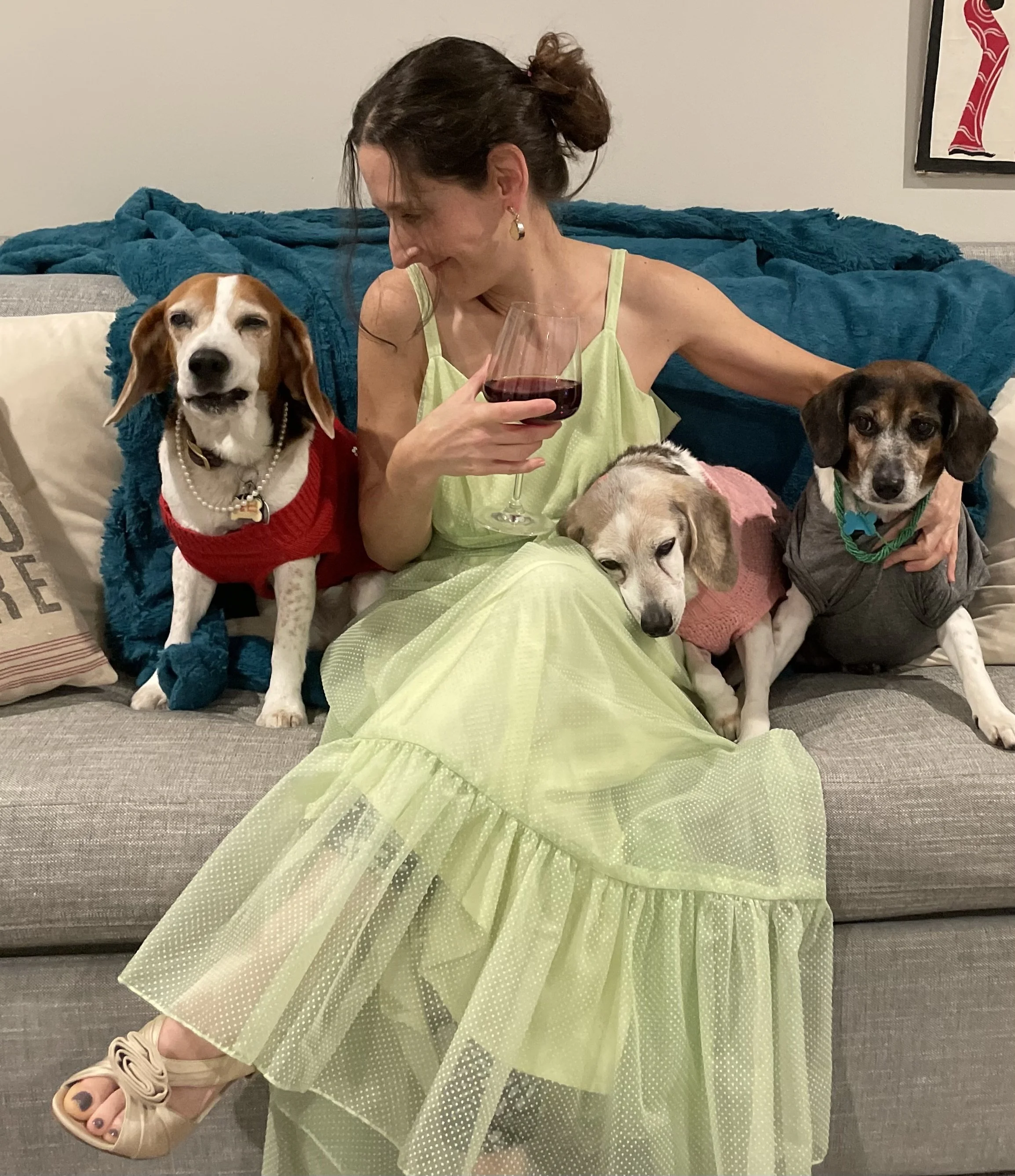 A woman, Katya Lidsky, with dark hair in a bun, wearing a yellow dress, surrounded by four dogs, sitting on a couch with a blue blanket and pillows in the background.