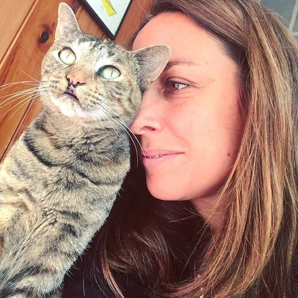 A woman with brown hair sitting close to a tabby cat, both facing the camera, indoors with wooden furniture in the background.