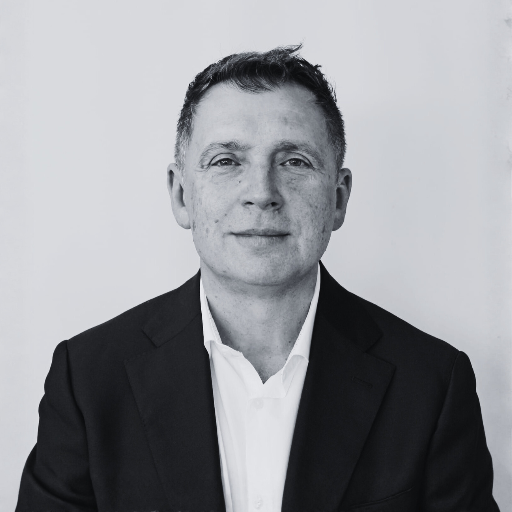 Black and white headshot of a man wearing a suit, smiling slightly, with a plain light background.