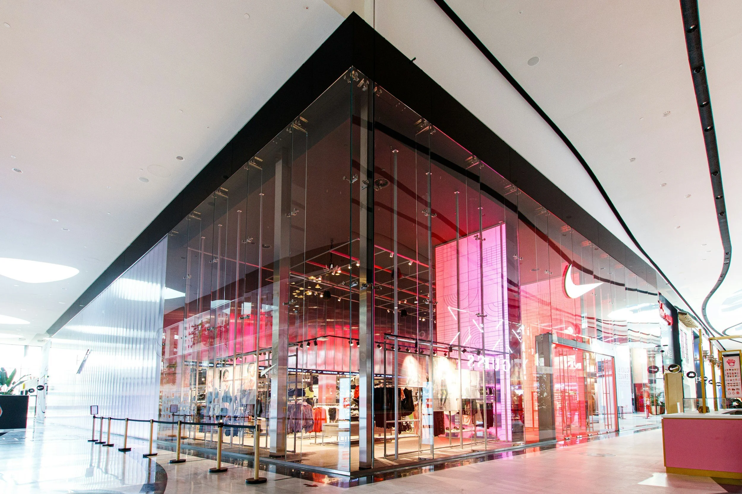 The image shows a modern Nike retail store inside a shopping mall with large glass walls, displaying clothing and mannequins, under pink and purple lighting.