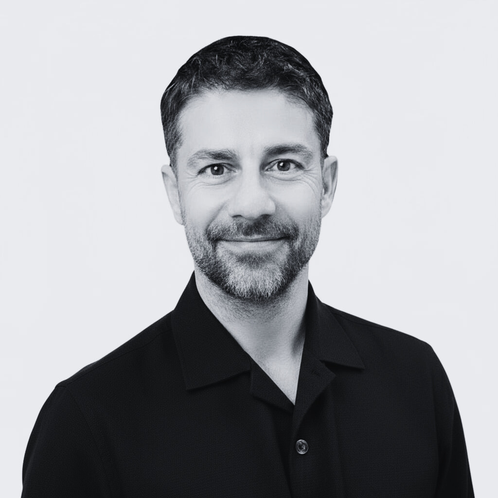 Black and white headshot of man with short dark hair, beard, wearing black collared shirt, smiling, against plain light background.