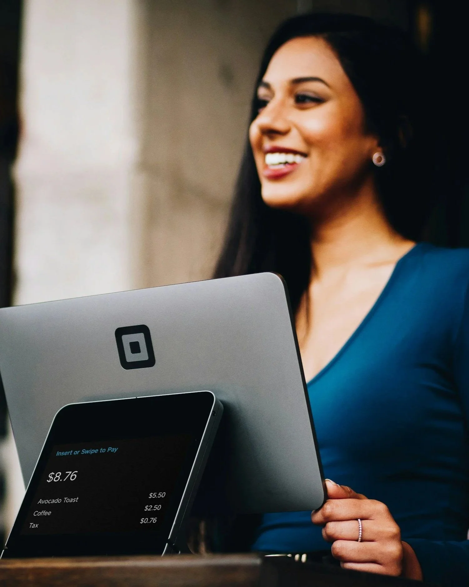 A smiling woman with long dark hair using a digital payment terminal, with an iPad and smartphone showing the total bill of $8.76 for avocado toast and coffee.