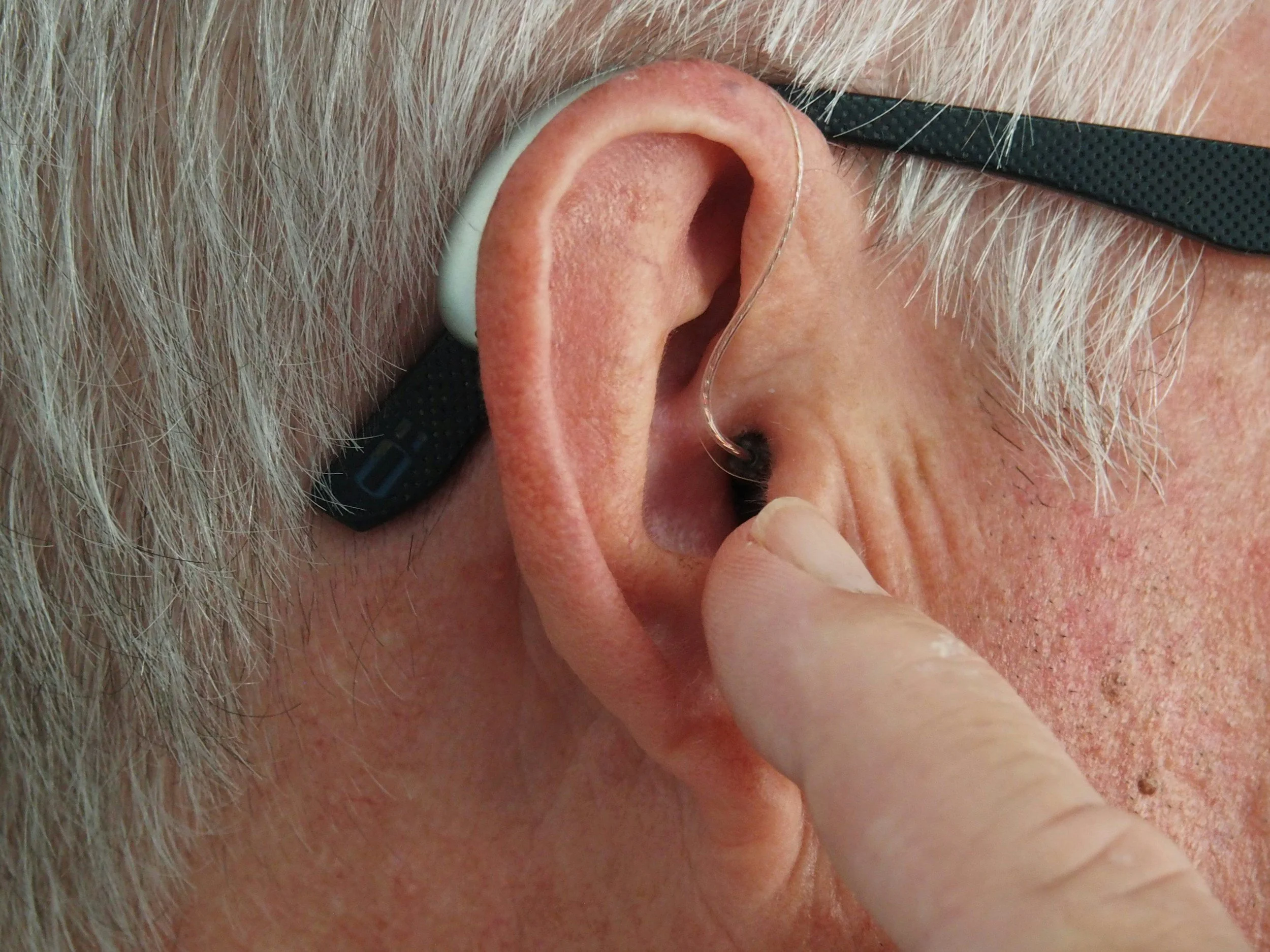 Close-up of an elderly man with white hair and glasses, demonstrating a person inserting a hearing aid into his ear.