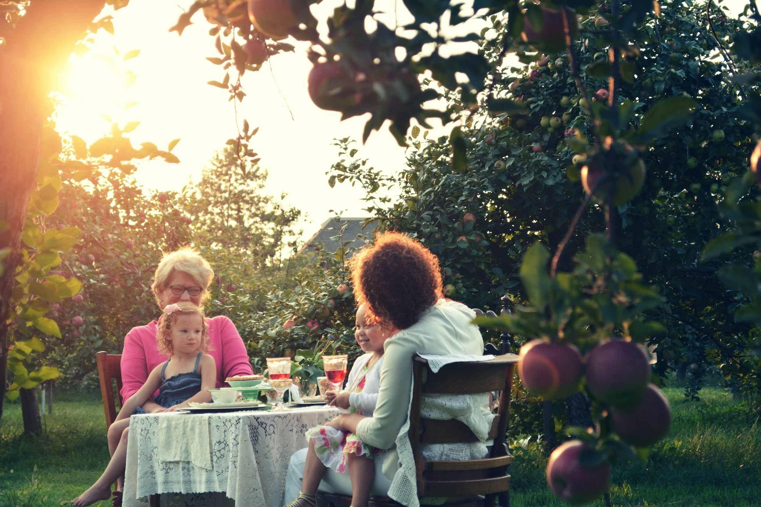 Four people, likely two adults and two children, are sitting at a table outdoors during sunset, surrounded by apple trees and fruit. They are enjoying drinks and food together.