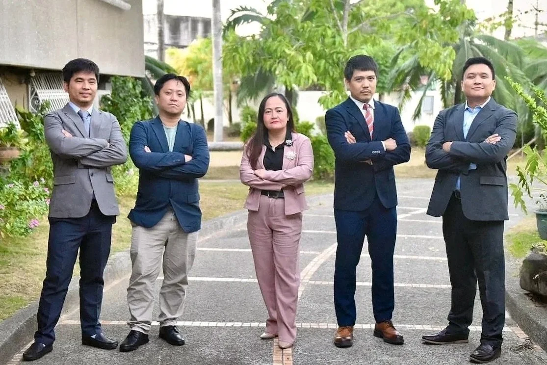 Five professionals standing outdoors in business attire with arms crossed, posing on a driveway with greenery in the background.