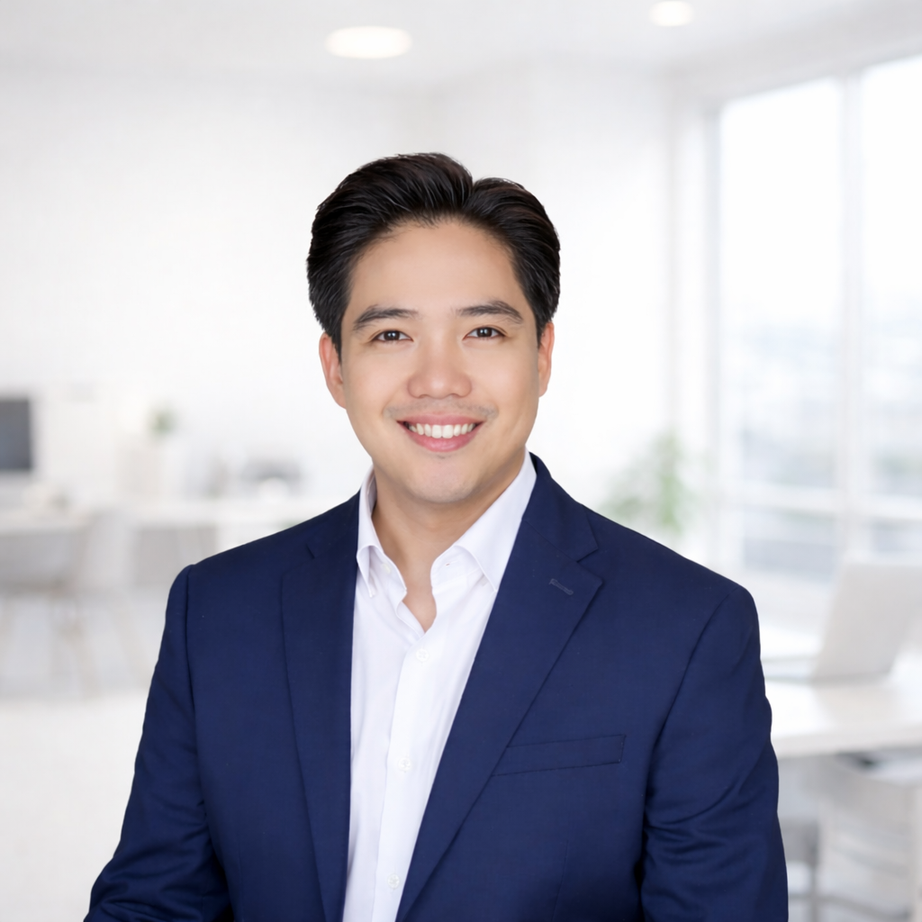 A smiling man in a business suit with a light blue shirt, standing in a modern office with large windows in the background.