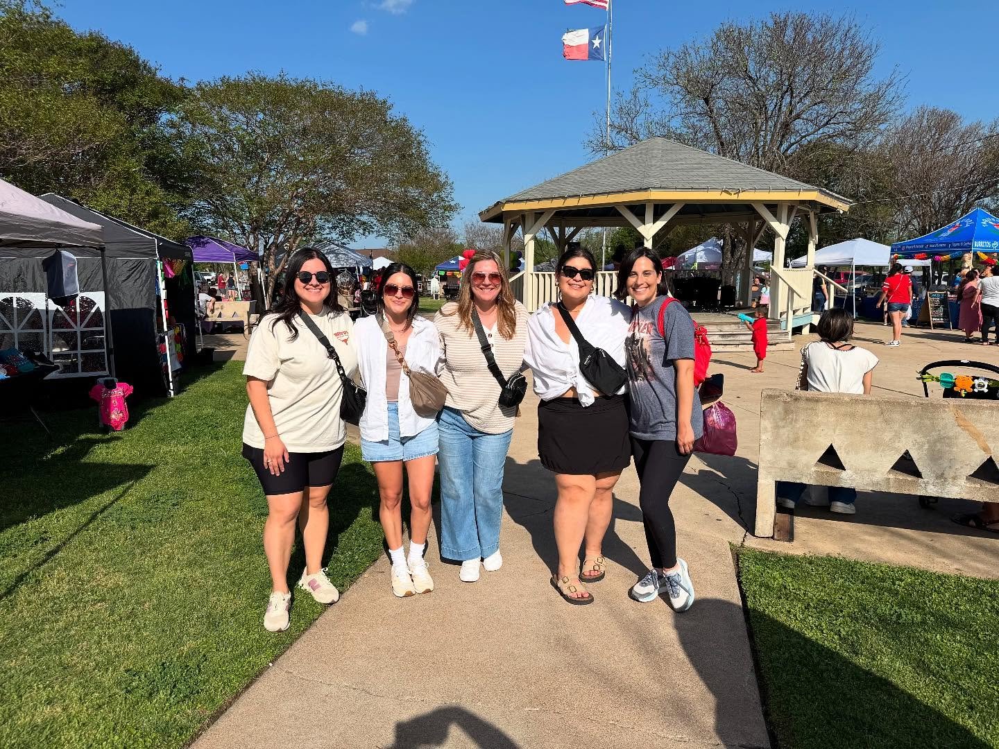 Five women standing together at an outdoor event on a sunny day, with tents, trees, and a pavilion in the background.