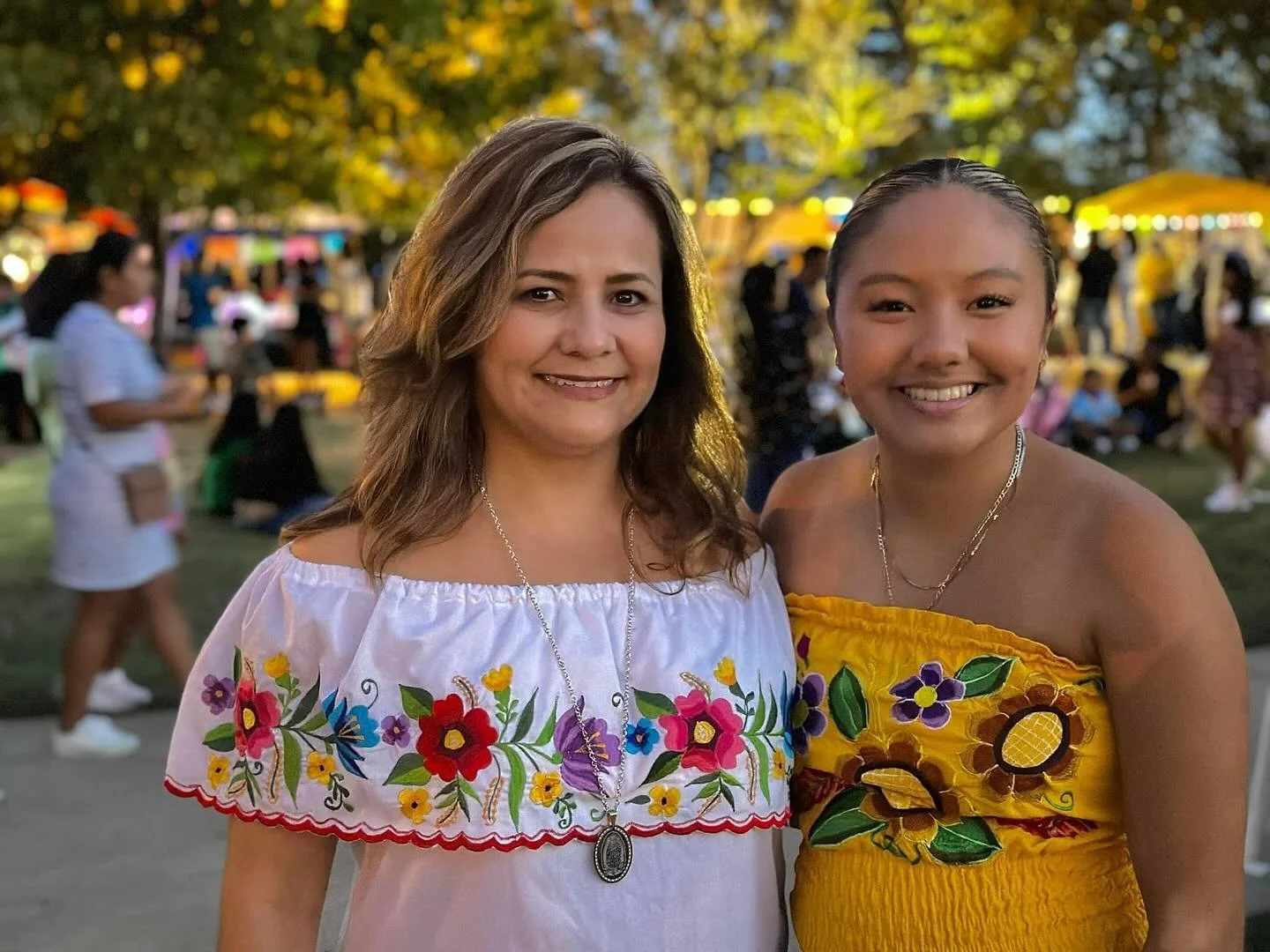 Two women smiling outdoors at a festival or fair, with trees, colorful booths, and other people in the background in the evening.