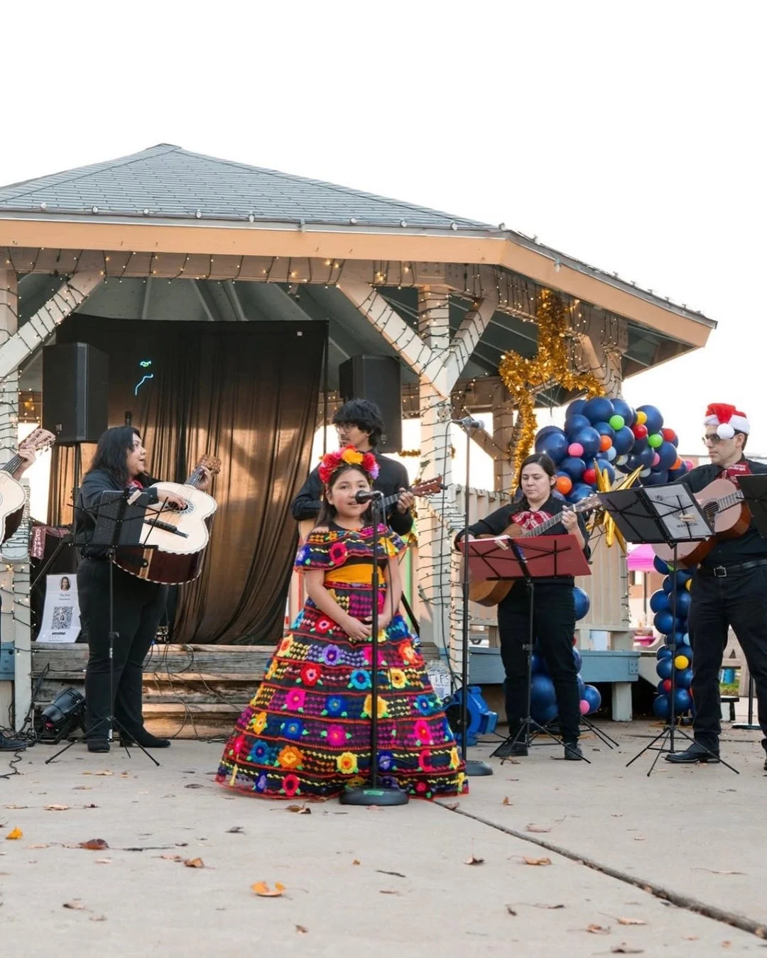 A group of musicians and a singer performing on an outdoor stage decorated with holiday ornaments, including balloons, tinsel, and a Christmas hat, with a young girl in traditional Mexican dress singing into a microphone.