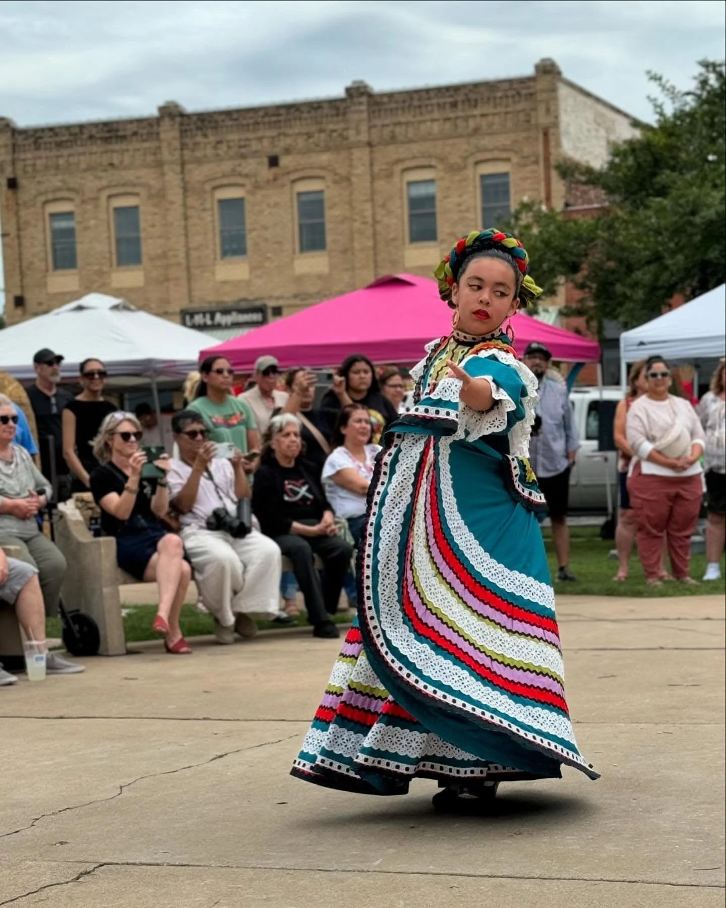 A woman dressed in a colorful traditional Mexican dress performs a dance at an outdoor event, watched by an audience seated on benches and standing, with tents and a brick building in the background.