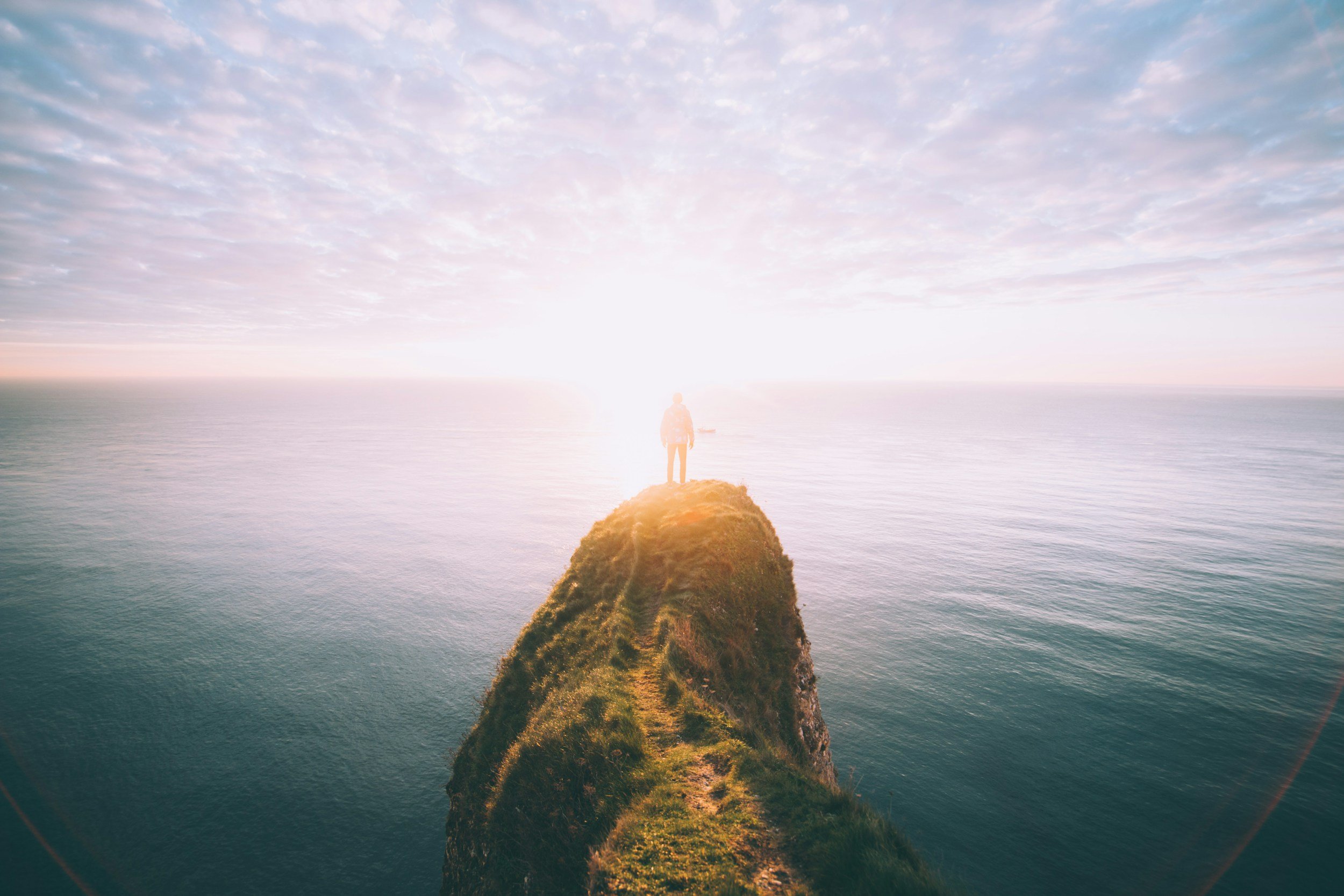 Person standing on a grassy cliff overlooking the ocean at sunrise or sunset with a bright sun in the distance
