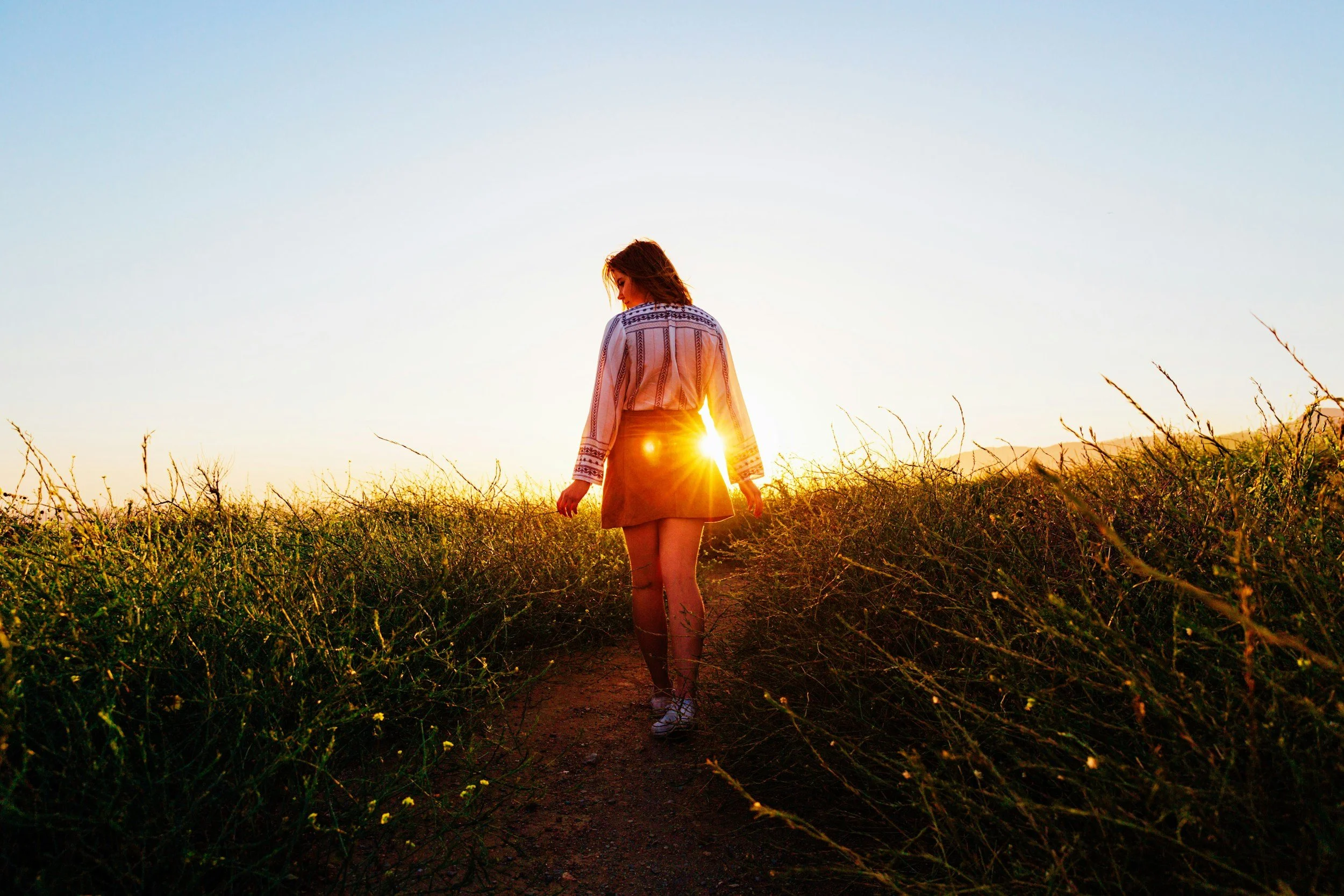 A woman walking on a trail through wild grass during sunset, with the sun setting behind her.