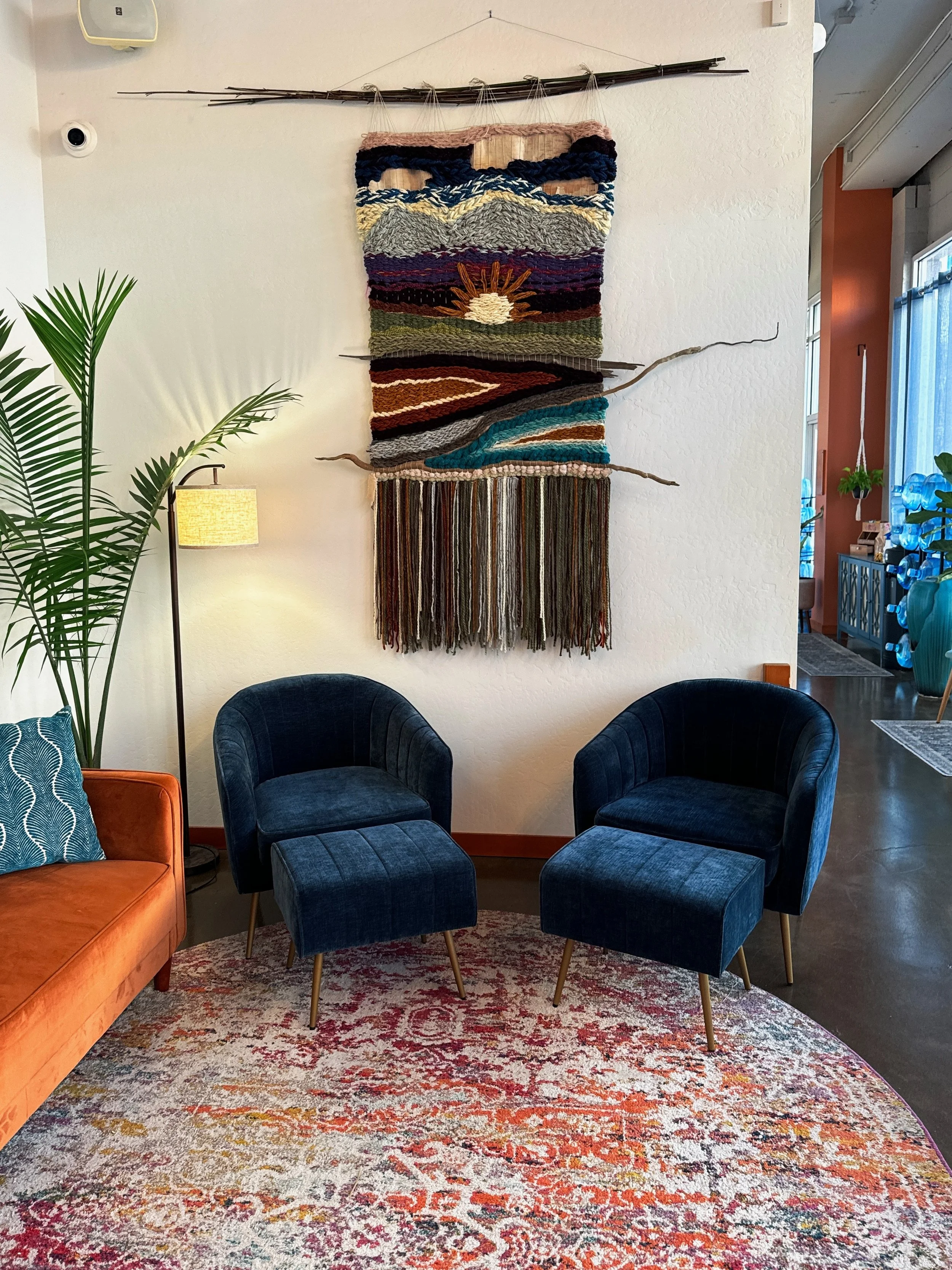 A cozy living room corner with two dark blue velvet chairs and a matching ottoman, a burnt orange sofa with decorative cushions, a tall green plant, and a colorful, textured wall hanging with horizontal stripes and a central sun motif, mounted on a white wall.