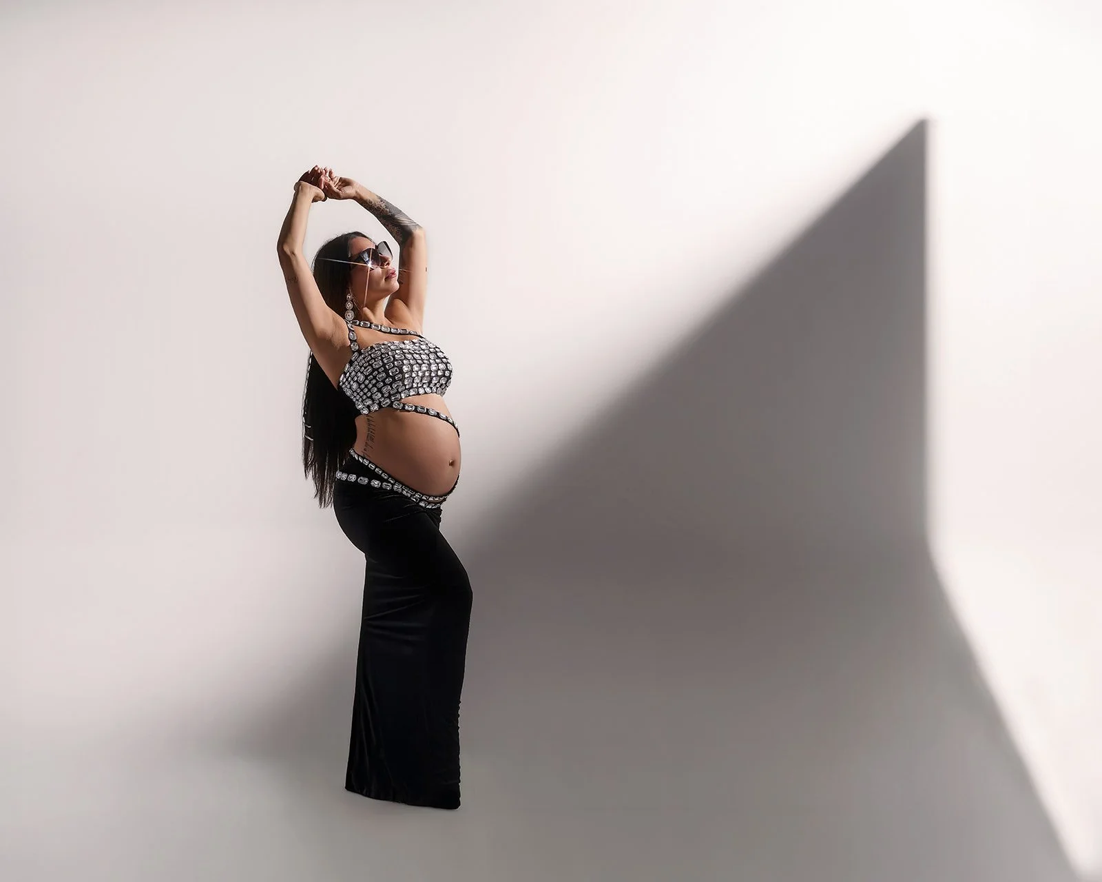 Pregnant woman in a jeweled top and black skirt posing against a white wall with geometric shadows in a Houston maternity studio.