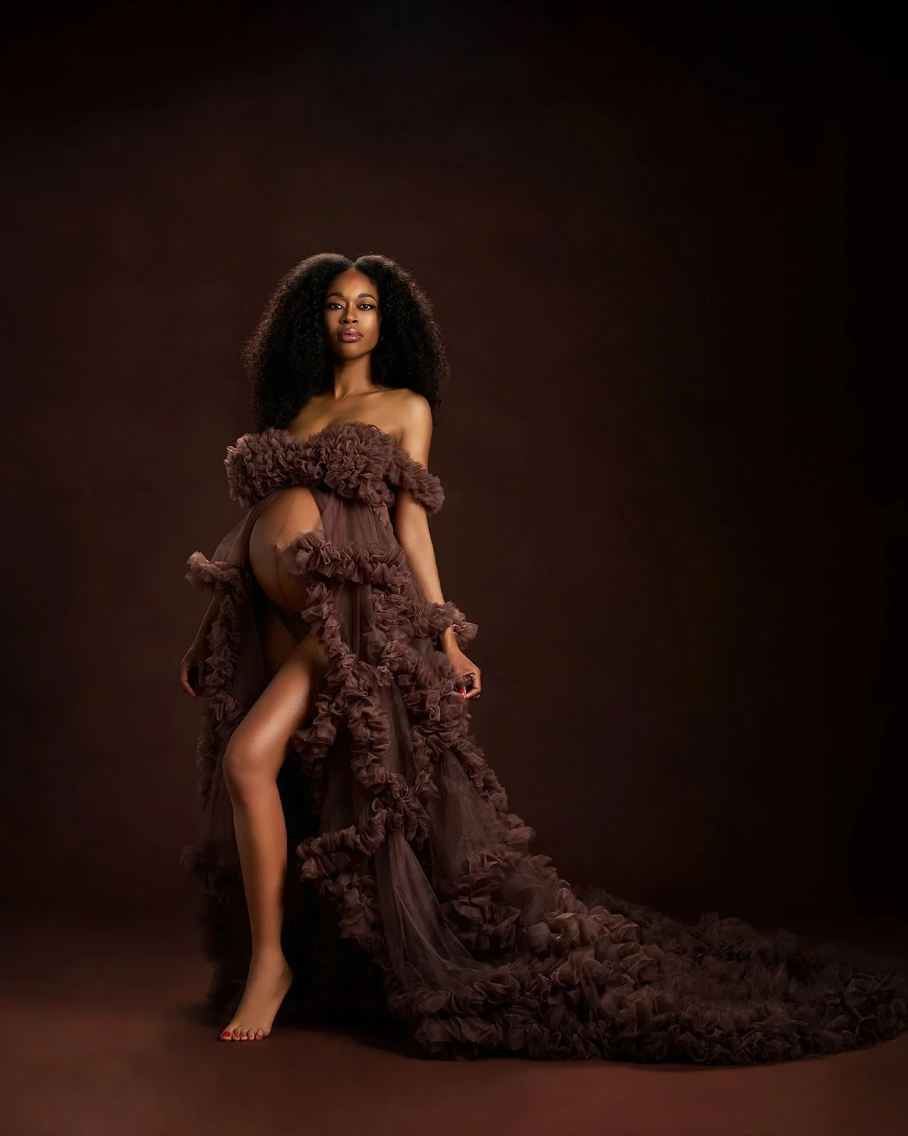 Full-length editorial maternity portrait featuring a flowing brown ruffled gown against a rich monochromatic background.