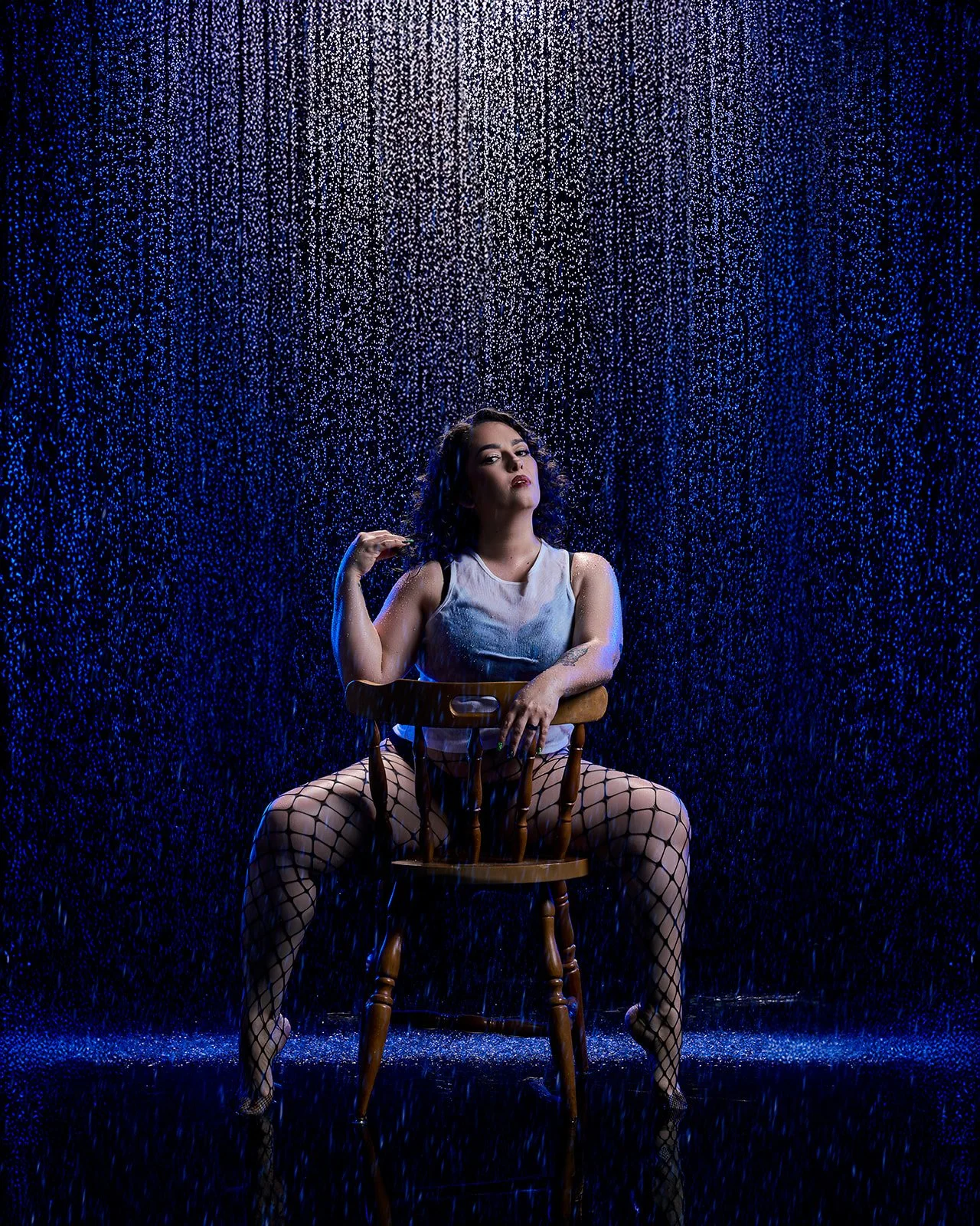 Woman posing backward on a wooden chair beneath falling water with dramatic blue studio lighting.