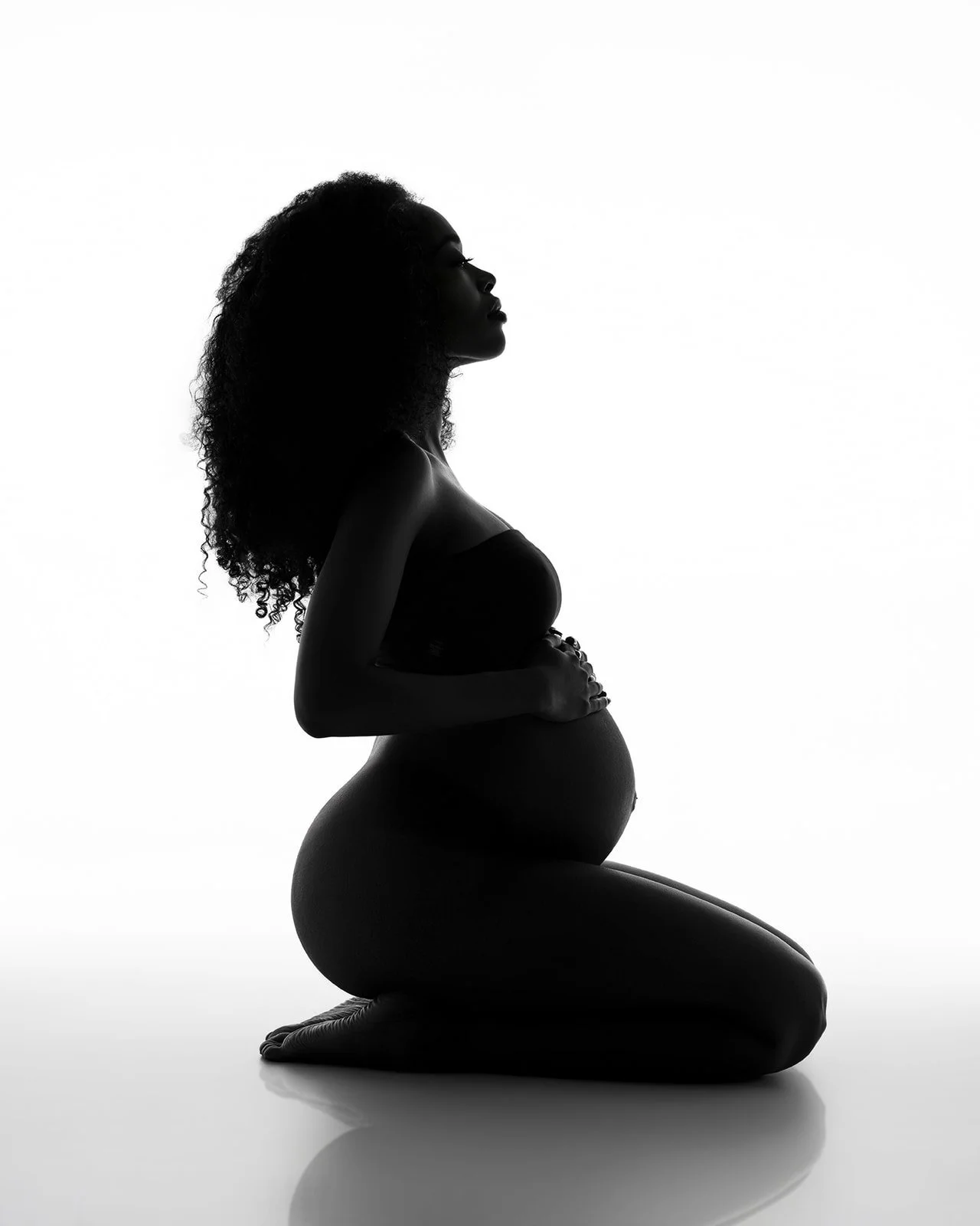 Kneeling pregnant woman photographed in silhouette against a bright white background in a Houston studio.