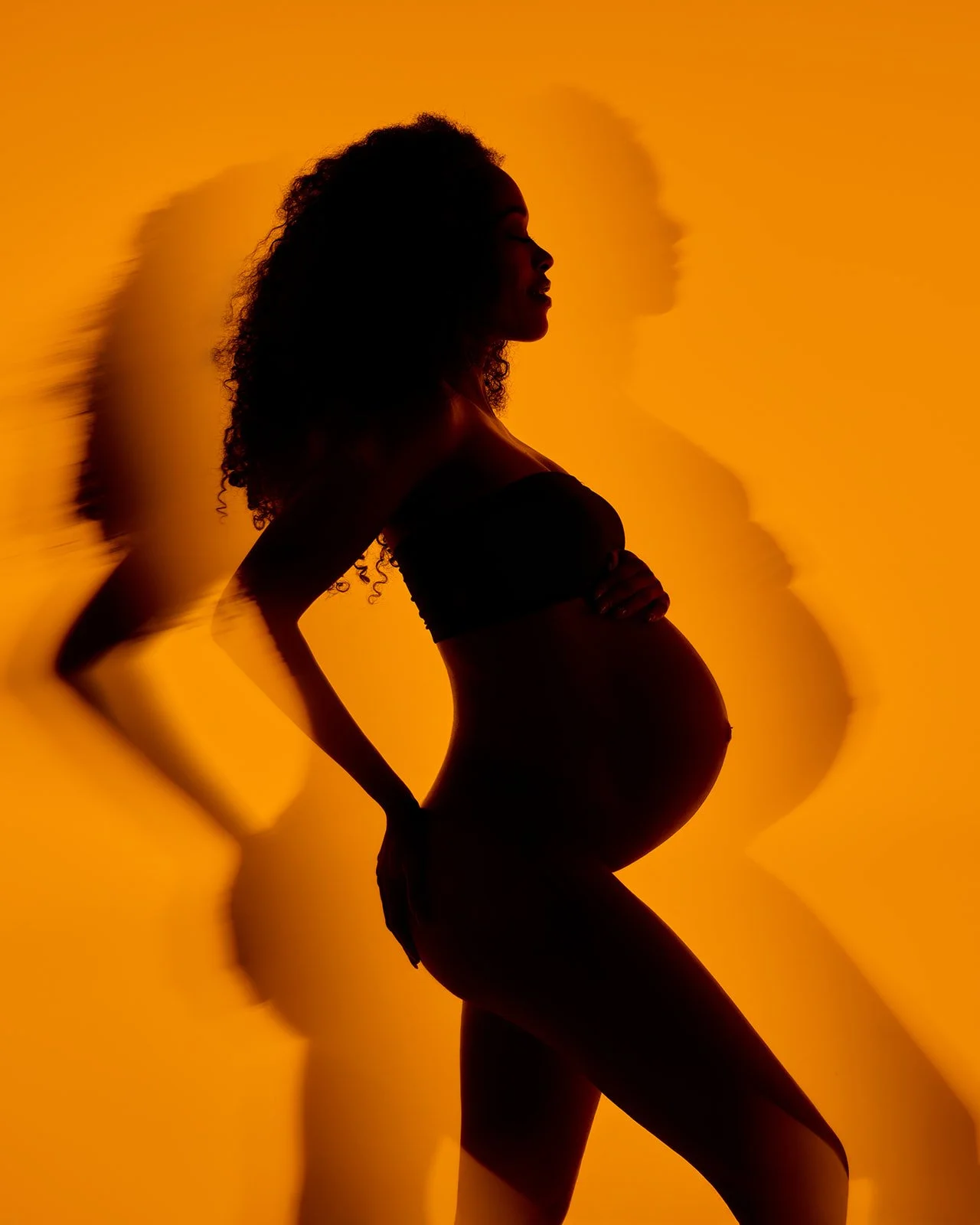 Artistic maternity silhouette with multiple shadows projected across a vivid orange studio background.