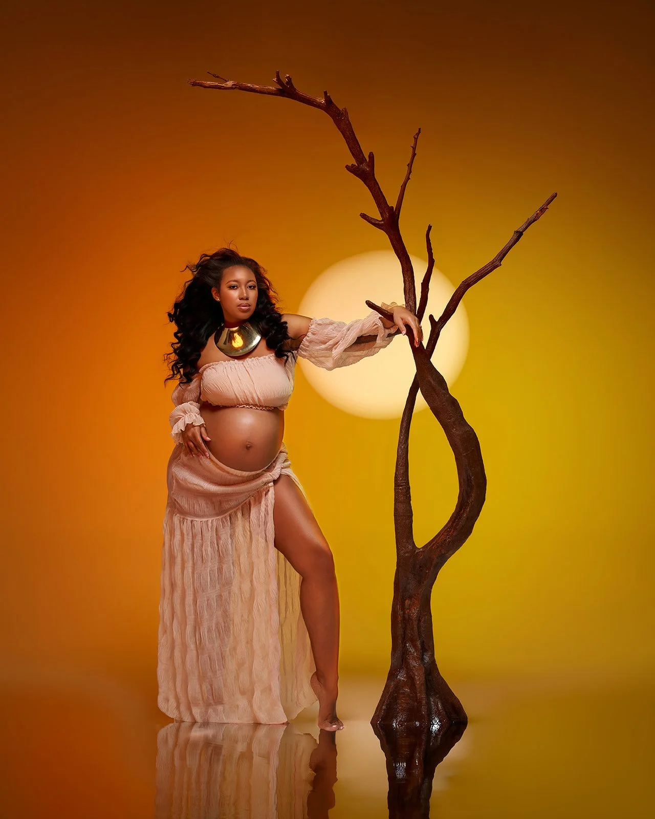 Pregnant woman posing beside a sculptural tree against a rich orange background with a glowing sun-inspired light.