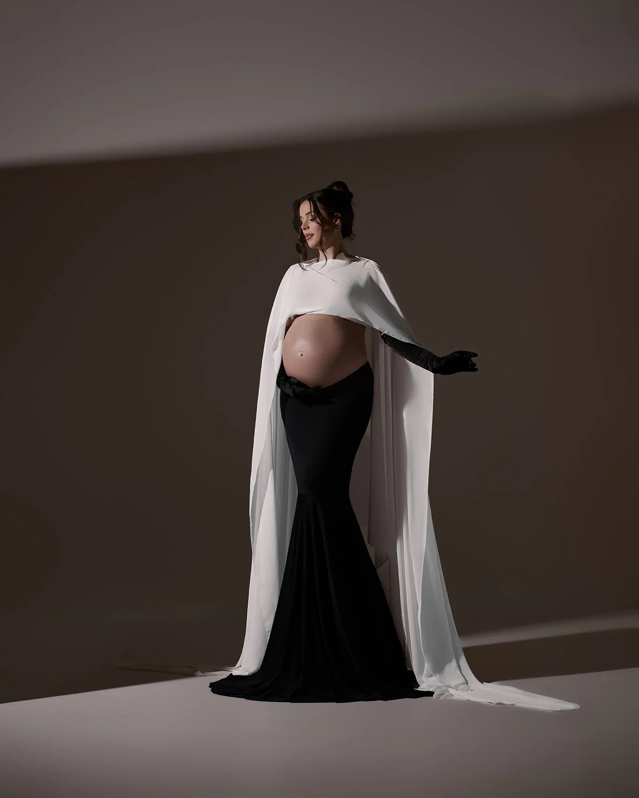 Pregnant woman wearing a black fitted skirt, white cape and long gloves in a dramatic editorial maternity portrait in Houston.
