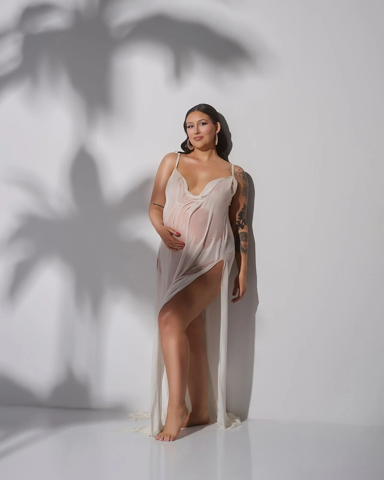 Pregnant woman wearing a sheer ivory dress posed against dramatic palm shadows in a modern Houston maternity studio portrait.