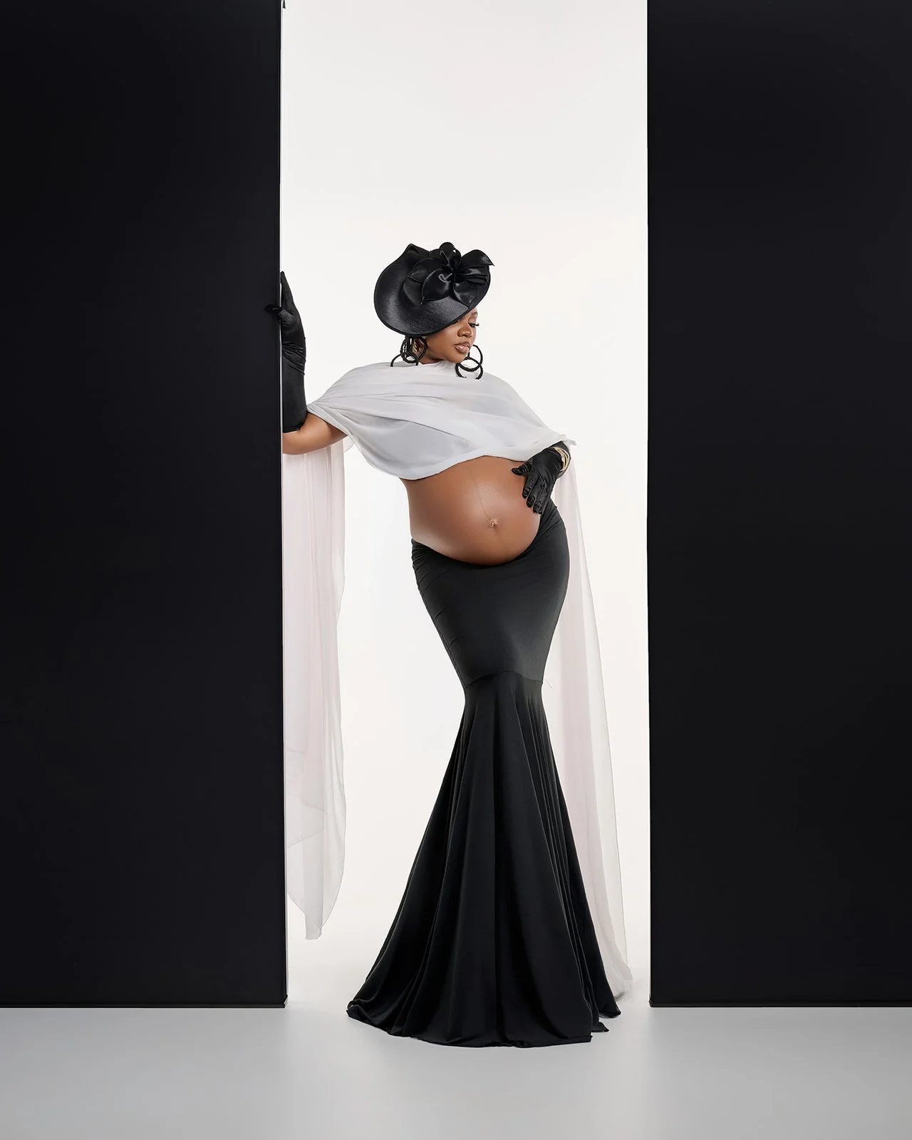 Pregnant woman wearing a black skirt, white draping, black gloves and statement hat in a fashion-inspired Houston maternity portrait.