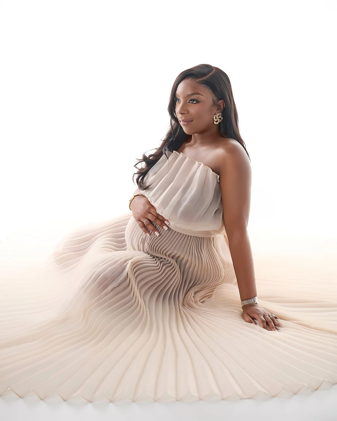 Pregnant woman seated in a flowing ivory pleated gown during an elegant studio maternity photography session in Houston.